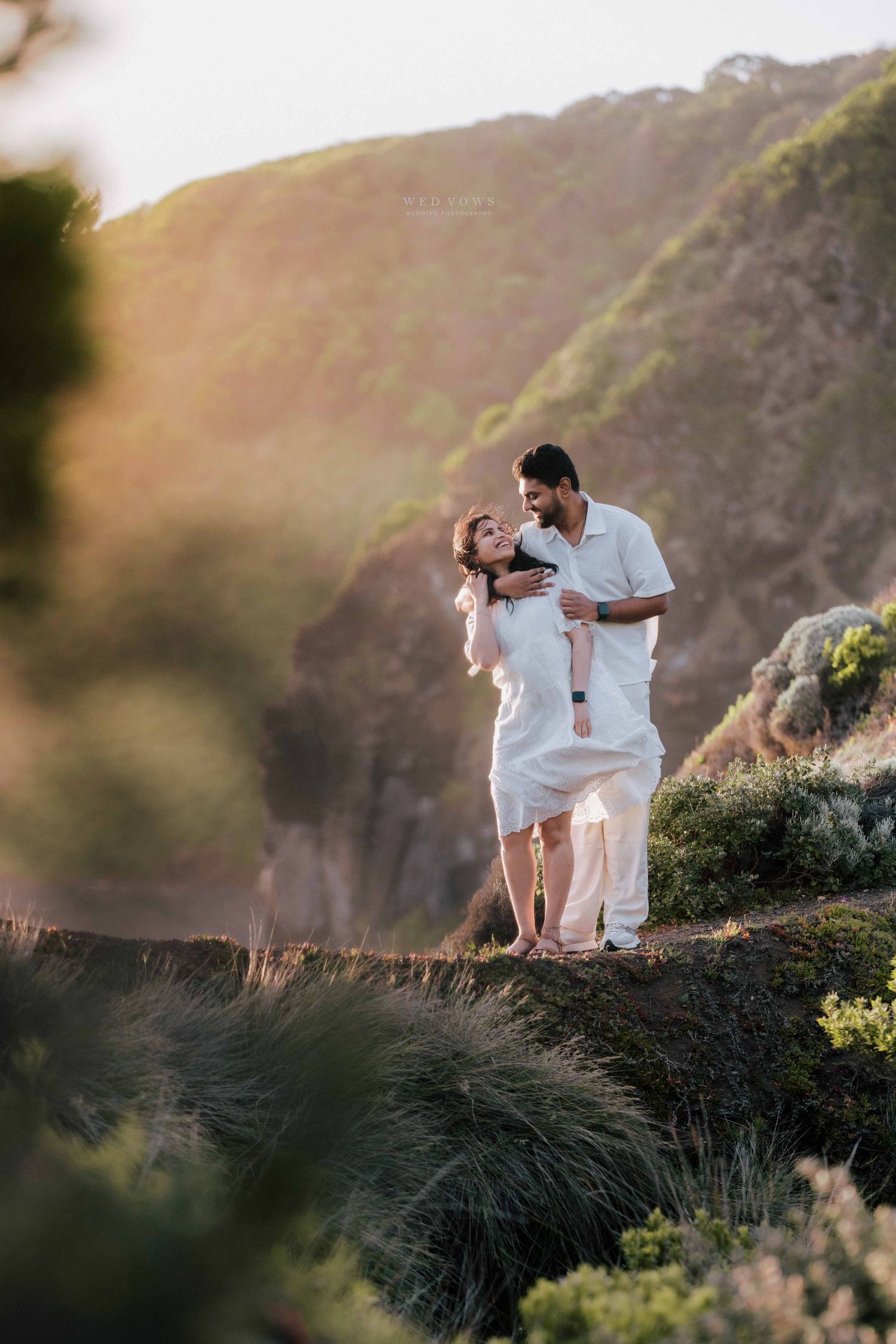Couple in white standing close together on scenic hillside with golden sunlight and greenery.