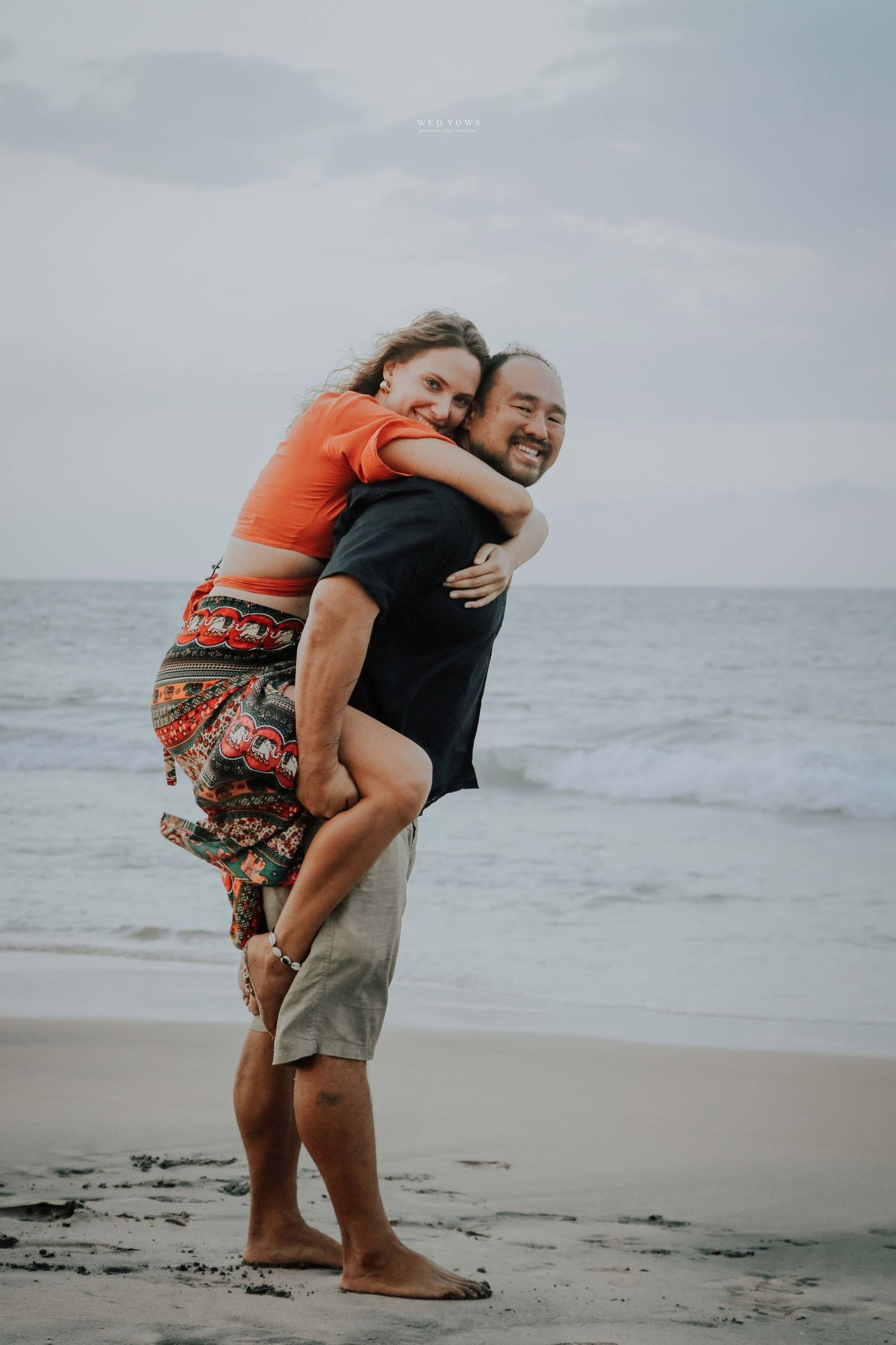 A man gives a woman a piggyback ride on a sandy beach, both smiling joyfully with ocean waves behind them.