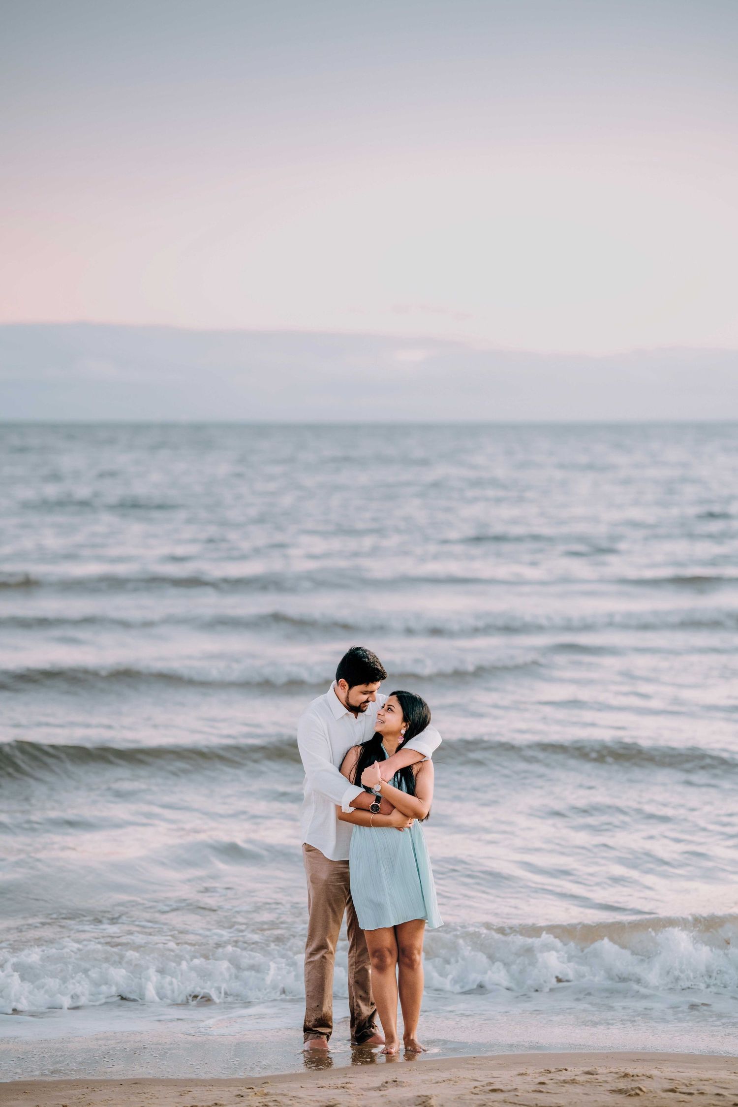 Indian couple embracing romantically on a sandy beach at sunset near ocean waves.