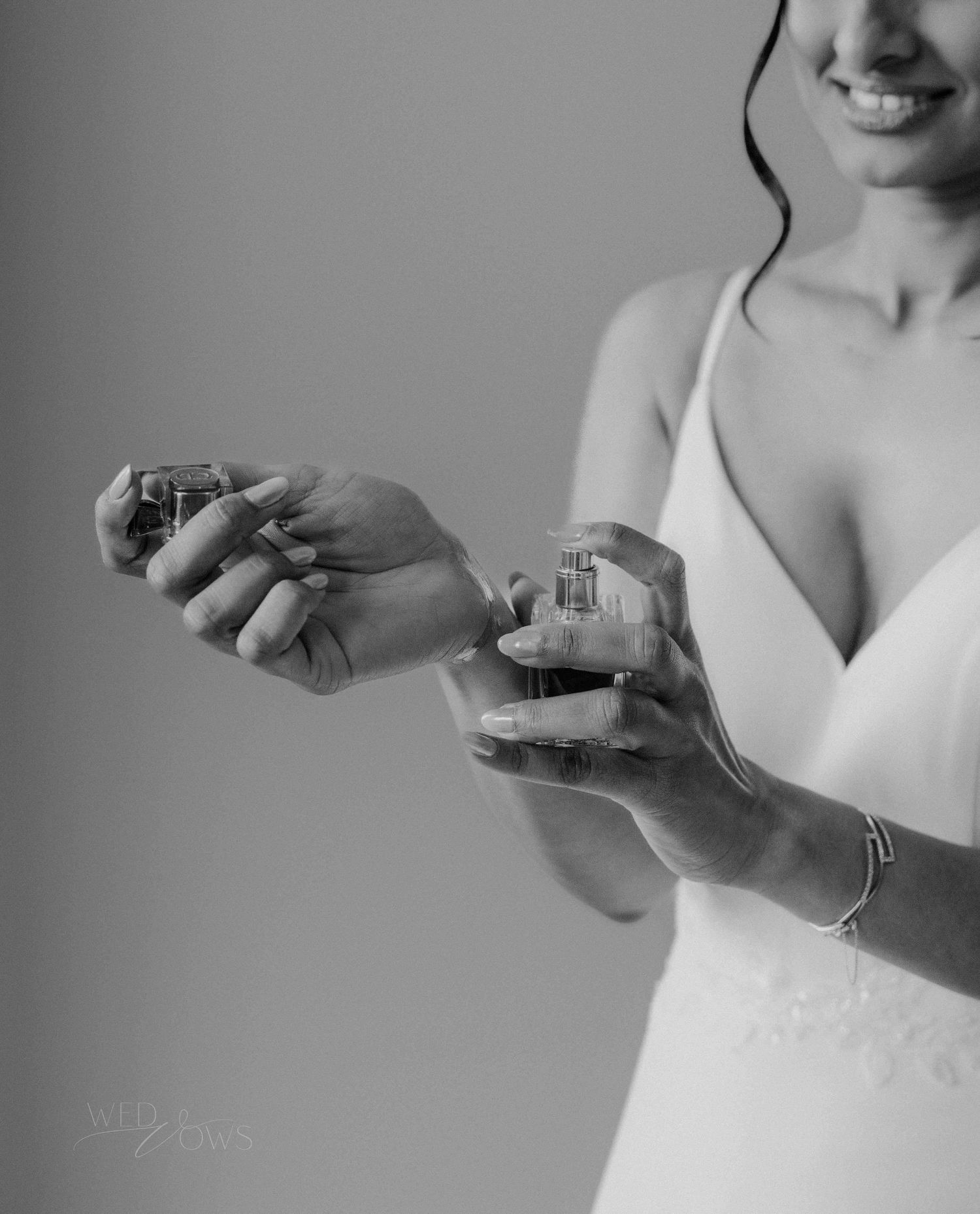 Black and white photo of bride in white gown holding two perfume bottles on her wedding day.