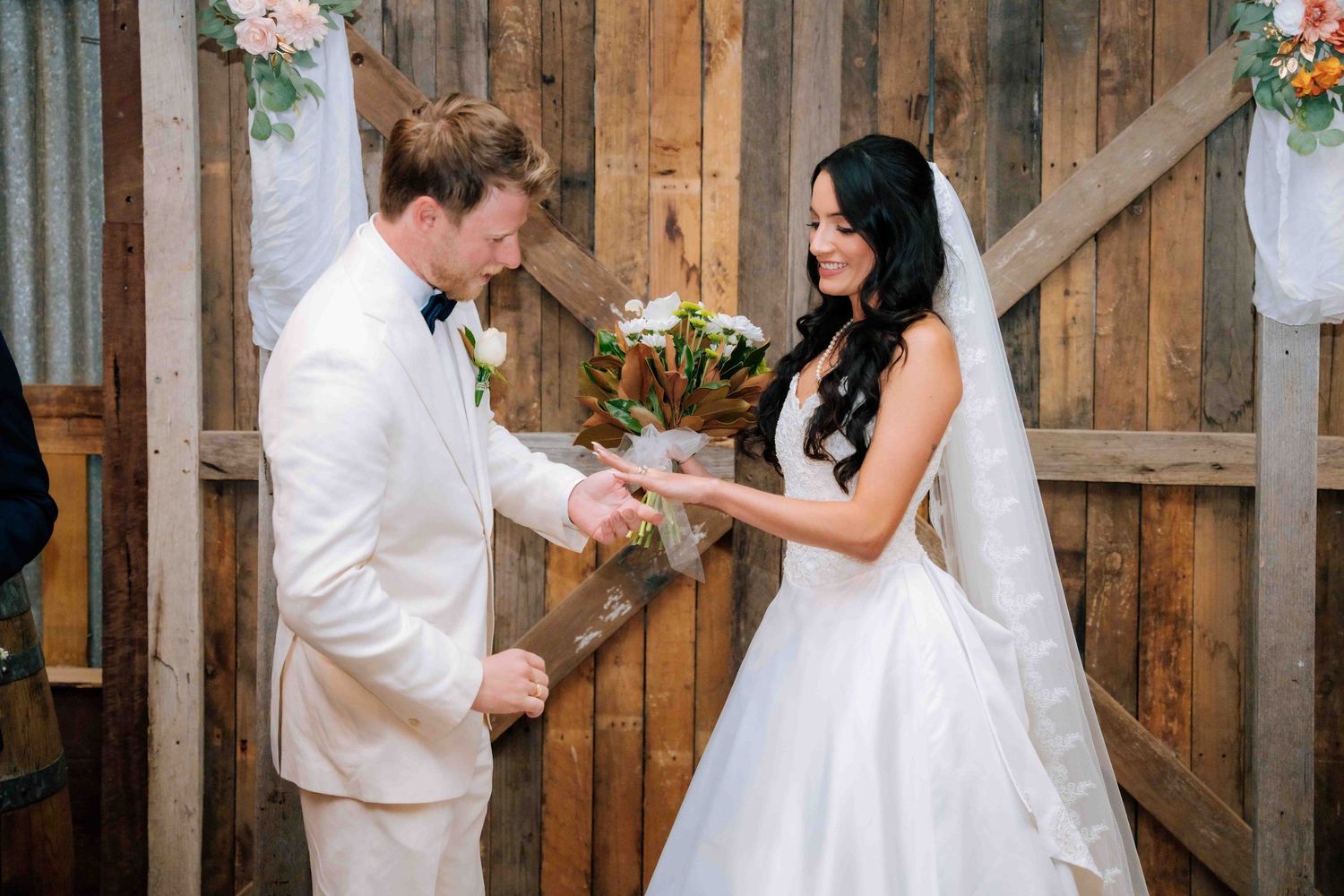 Bride and groom exchanging rings during rustic barn wedding ceremony, bride in white gown with veil holding bouquet.