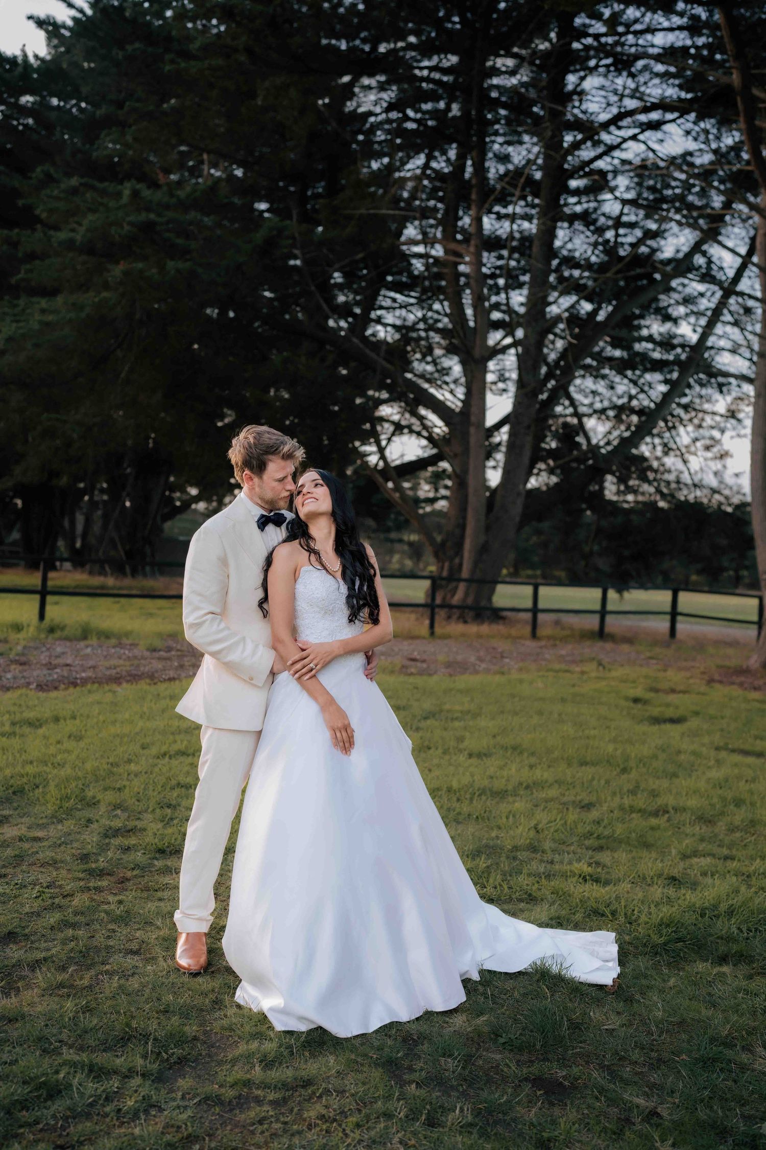 Bride and groom share a romantic embrace at golden hour in a lush green park with tall trees.