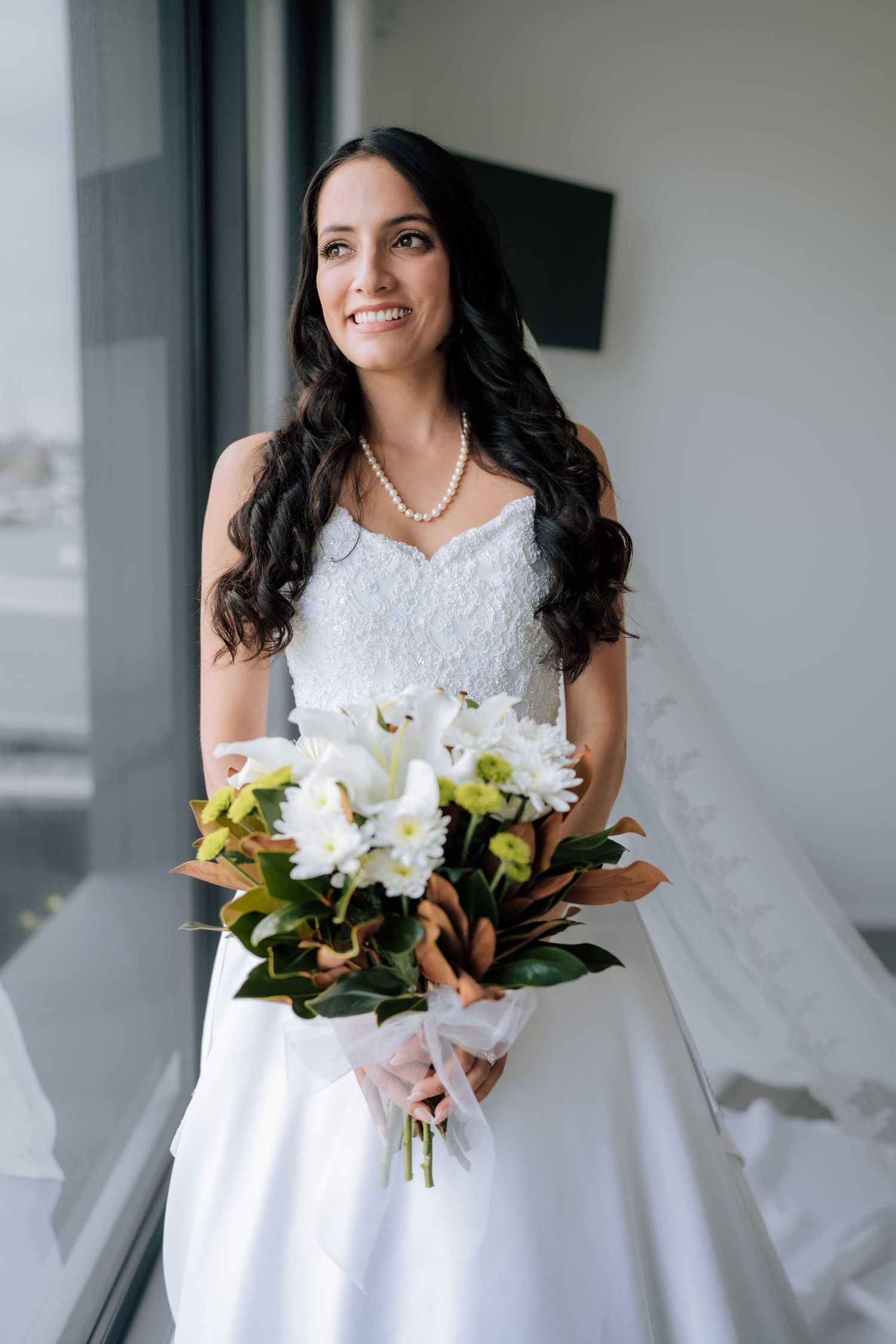 Smiling dark-haired bride in strapless lace gown holds white floral bouquet and wears pearl necklace indoors.