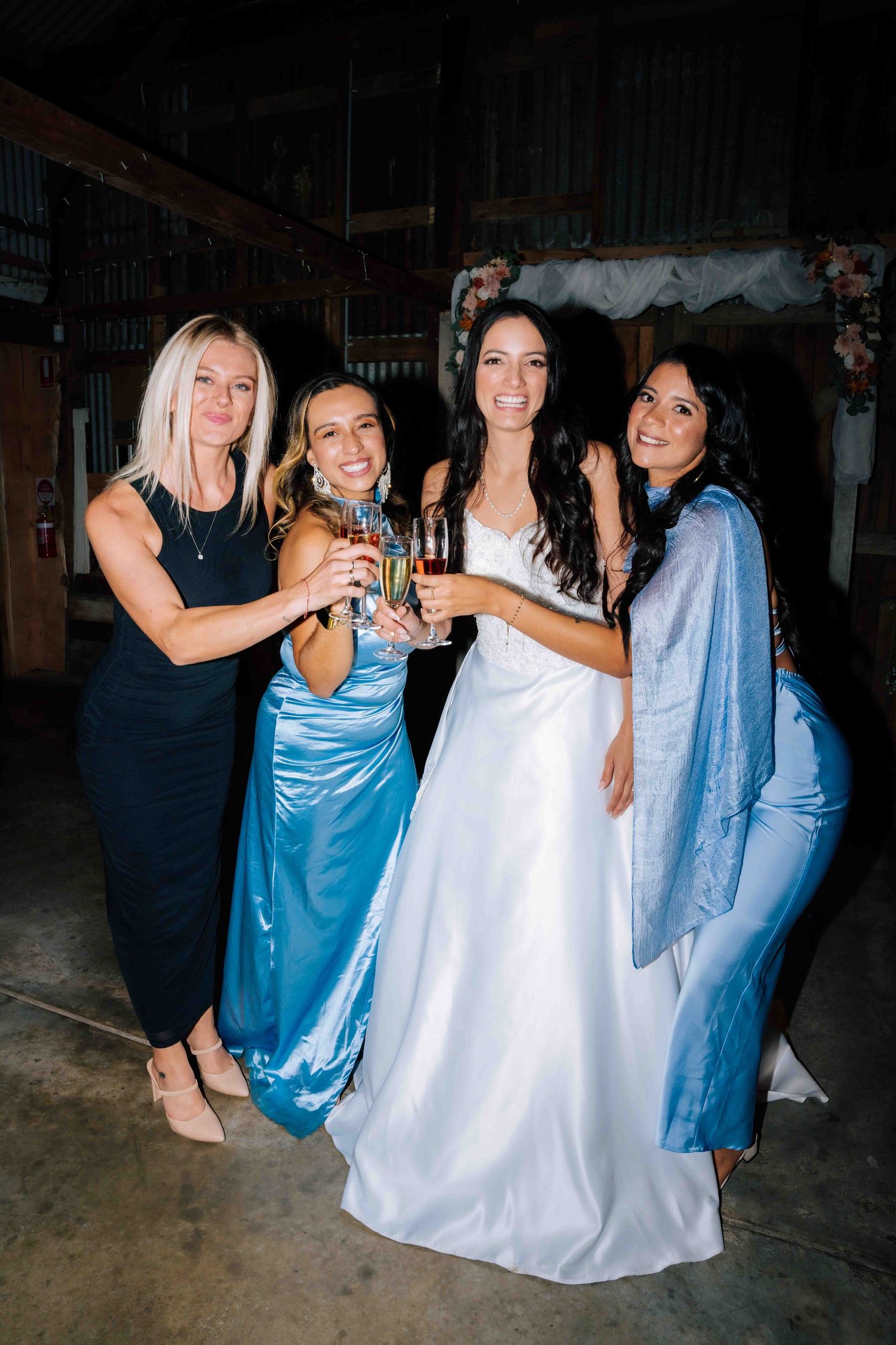 Bride in white gown toasting champagne with three guests in blue dresses at a rustic wedding venue.