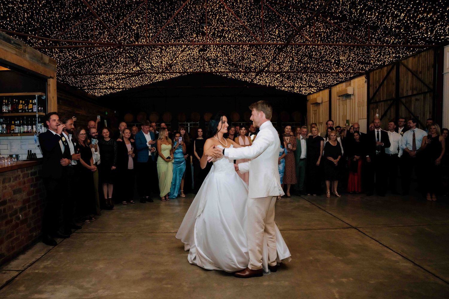 Bride and groom share first dance under fairy lights in rustic barn venue.