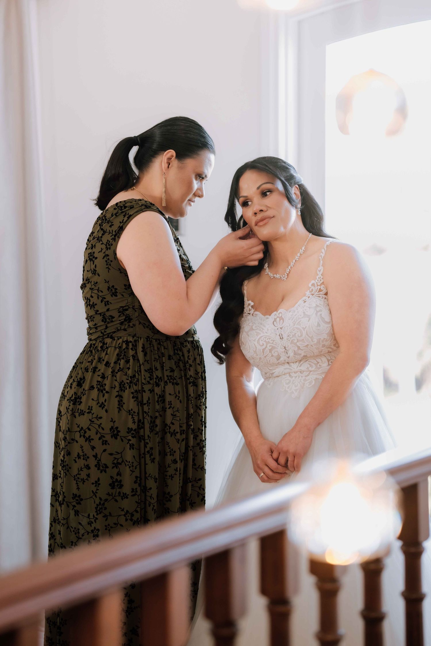 A woman in a floral dress helps a bride in a lace gown with her earring on a staircase.