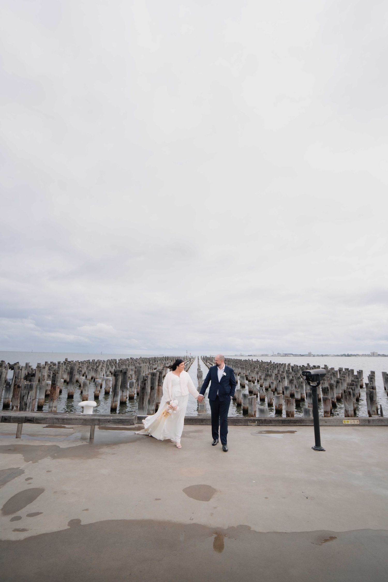 Bride and groom holding hands walking along a seaside pier with wooden pylons under an overcast sky.