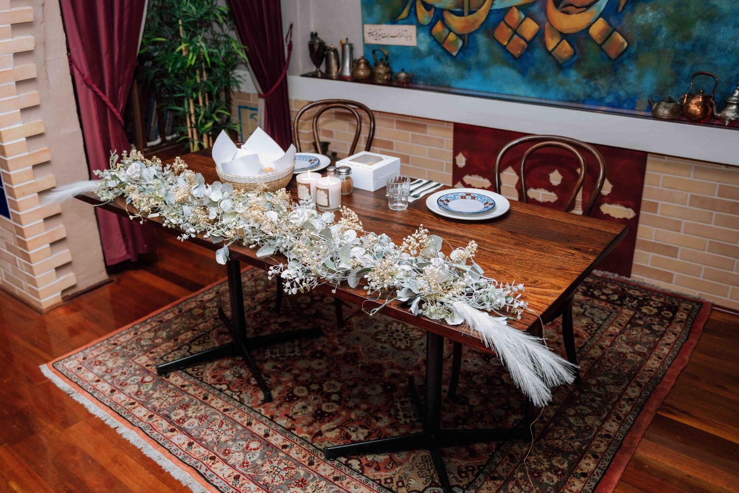 Rustic wooden wedding head table decorated with white floral garland runner and candles on patterned rug.