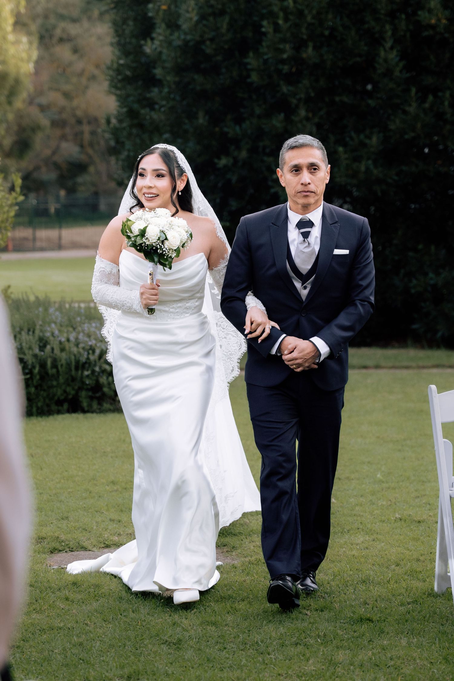 Bride in white gown with veil walking down aisle with escort at outdoor garden wedding.