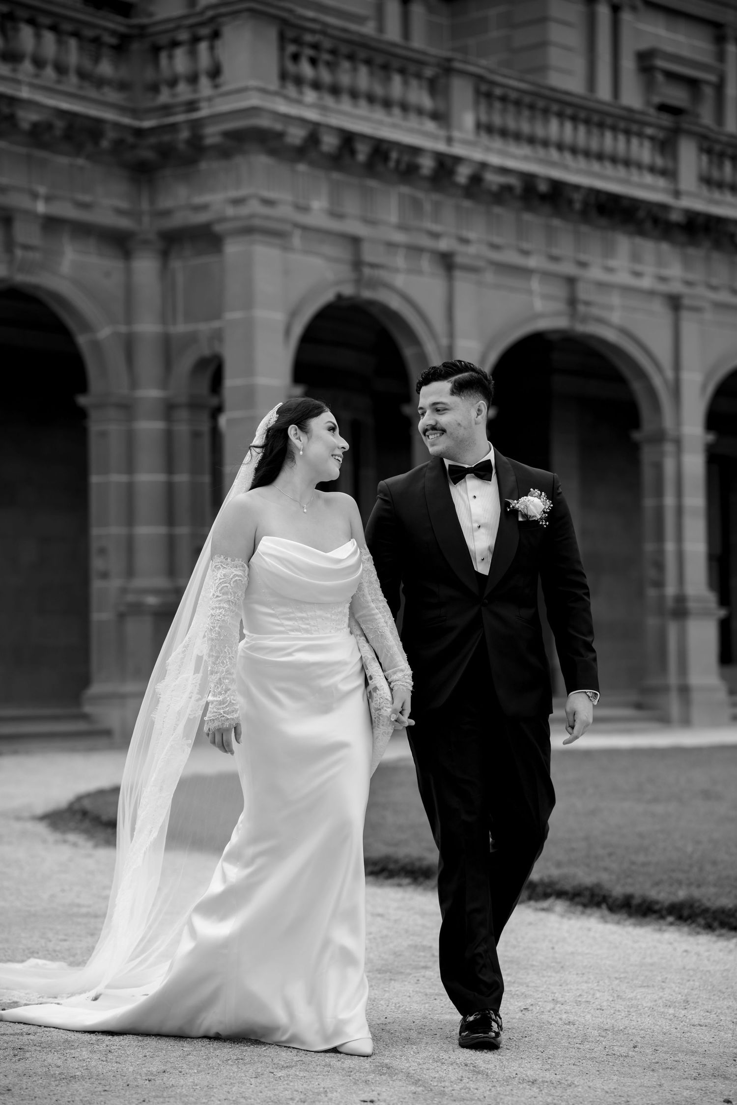 Black and white photo of a bride and groom walking hand in hand, smiling at each other outside a grand building.