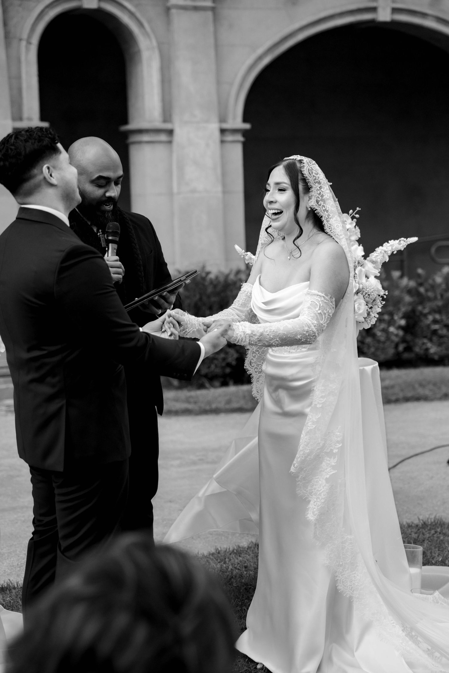 Black and white photo of joyful bride laughing while exchanging vows with groom at outdoor ceremony.