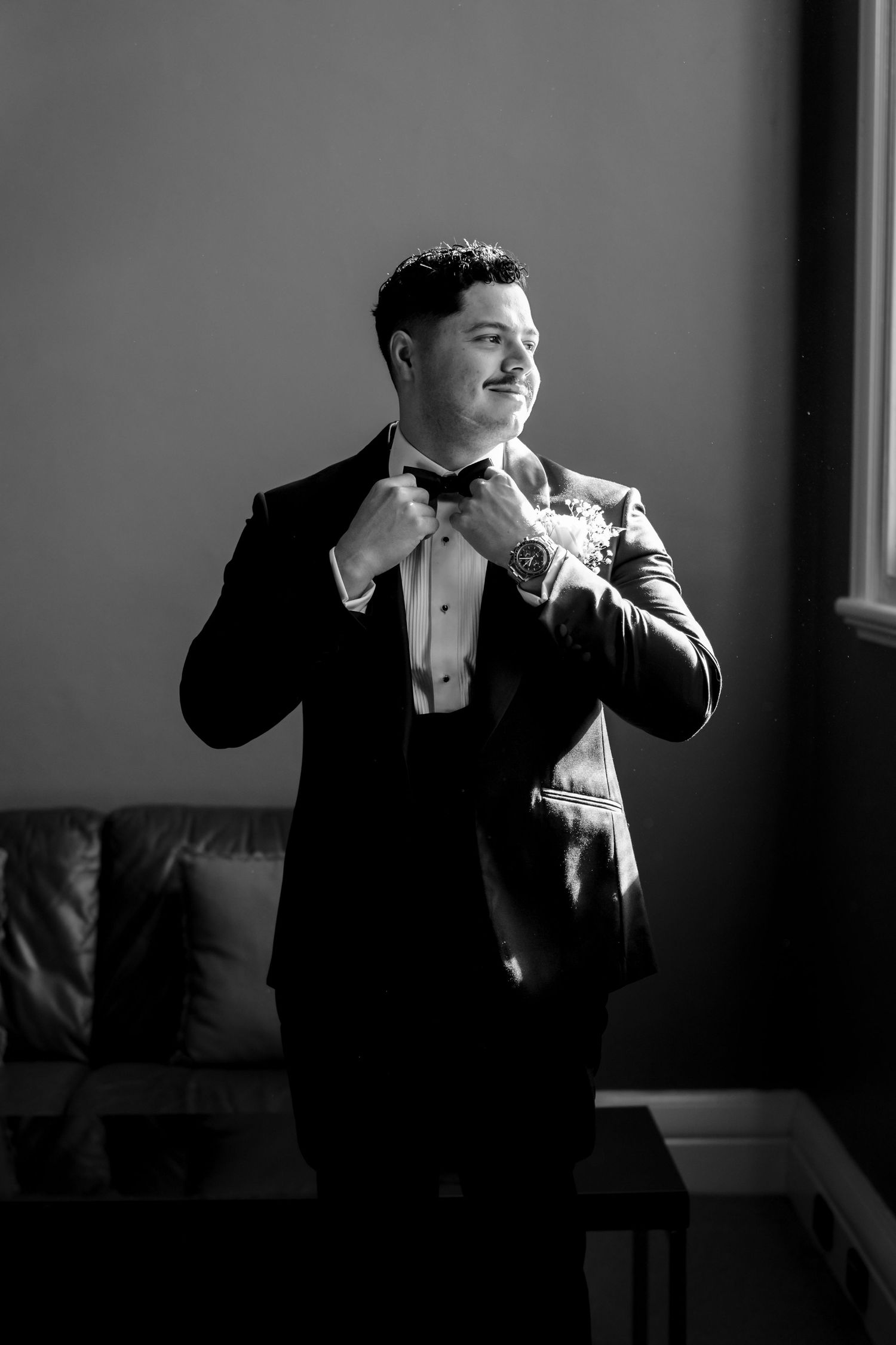 Groom in black tuxedo adjusting bow tie in black and white portrait near window.