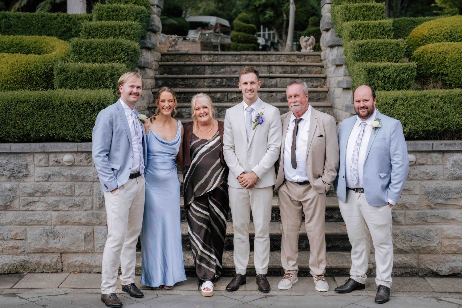 Groom stands with family members in formal attire on stone steps at wedding venue.