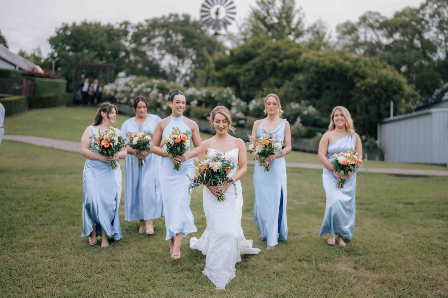 Bride and five bridesmaids in light blue dresses walk together on grass with windmill and rustic venue in background.
