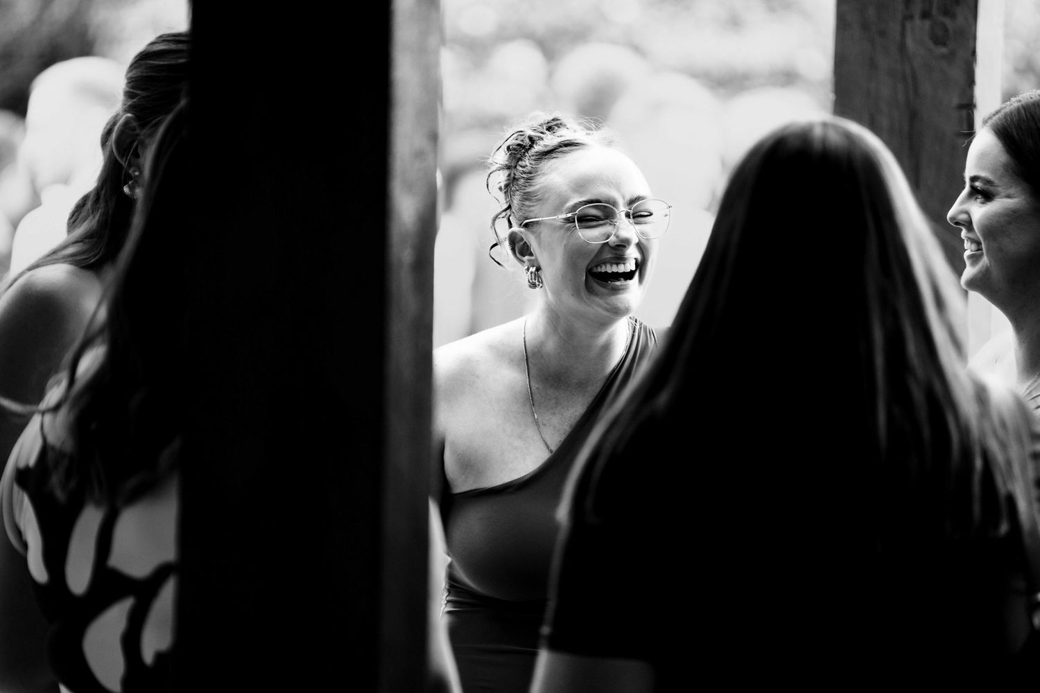Black and white photo of a laughing woman in a sleeveless dress surrounded by friends at a wedding celebration.