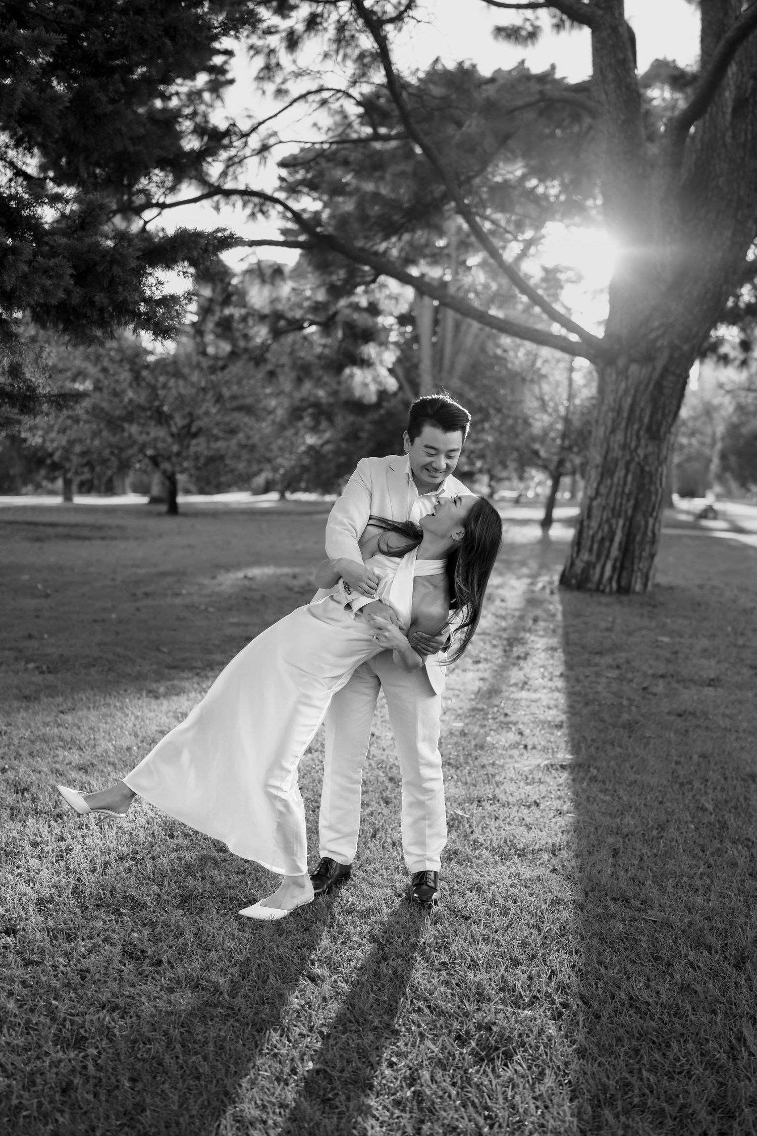 Black and white photo of a couple in wedding attire, groom dipping bride in a sunlit park.