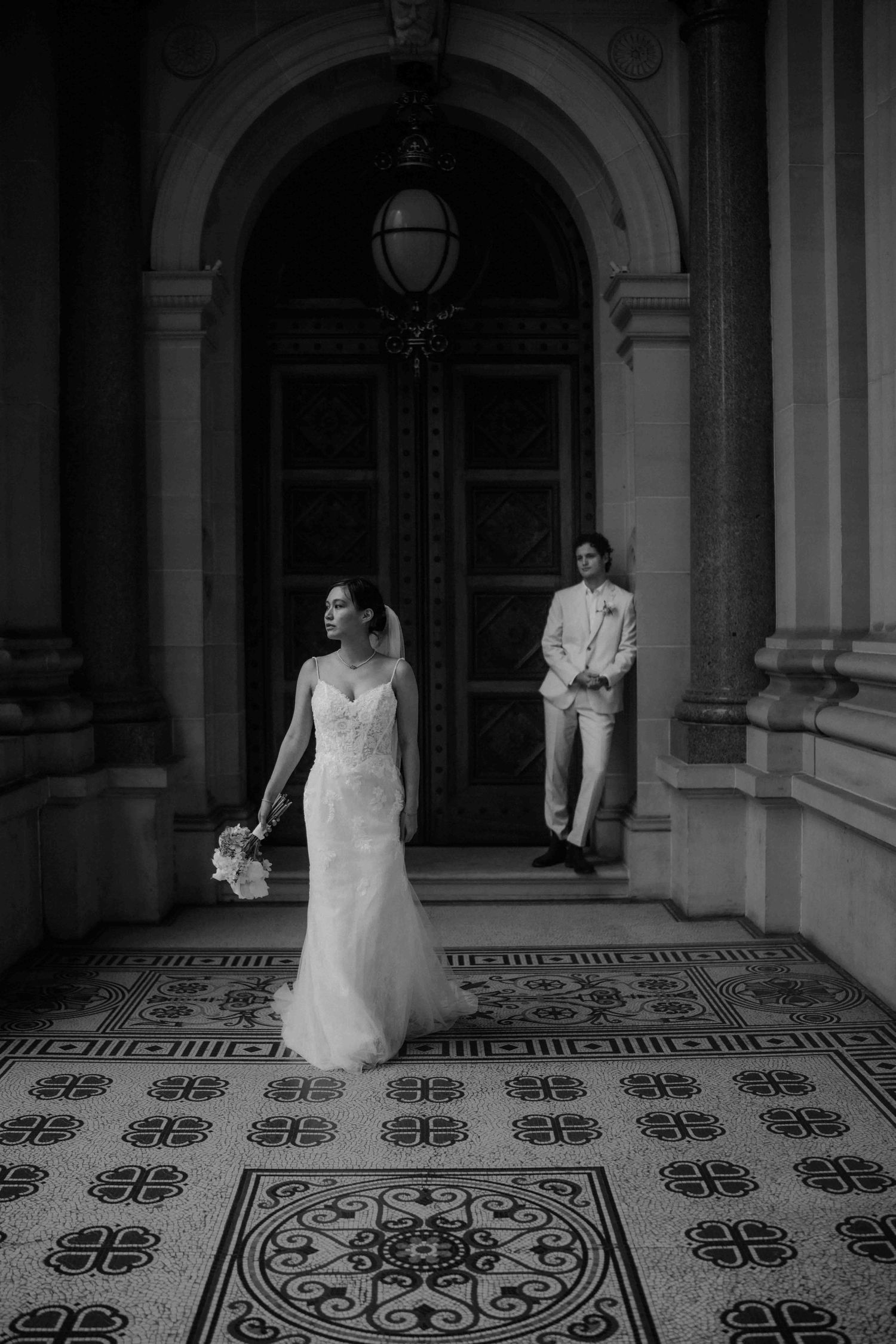 Bride and groom posing in elegant arched doorway with ornate tiled floor in black and white.