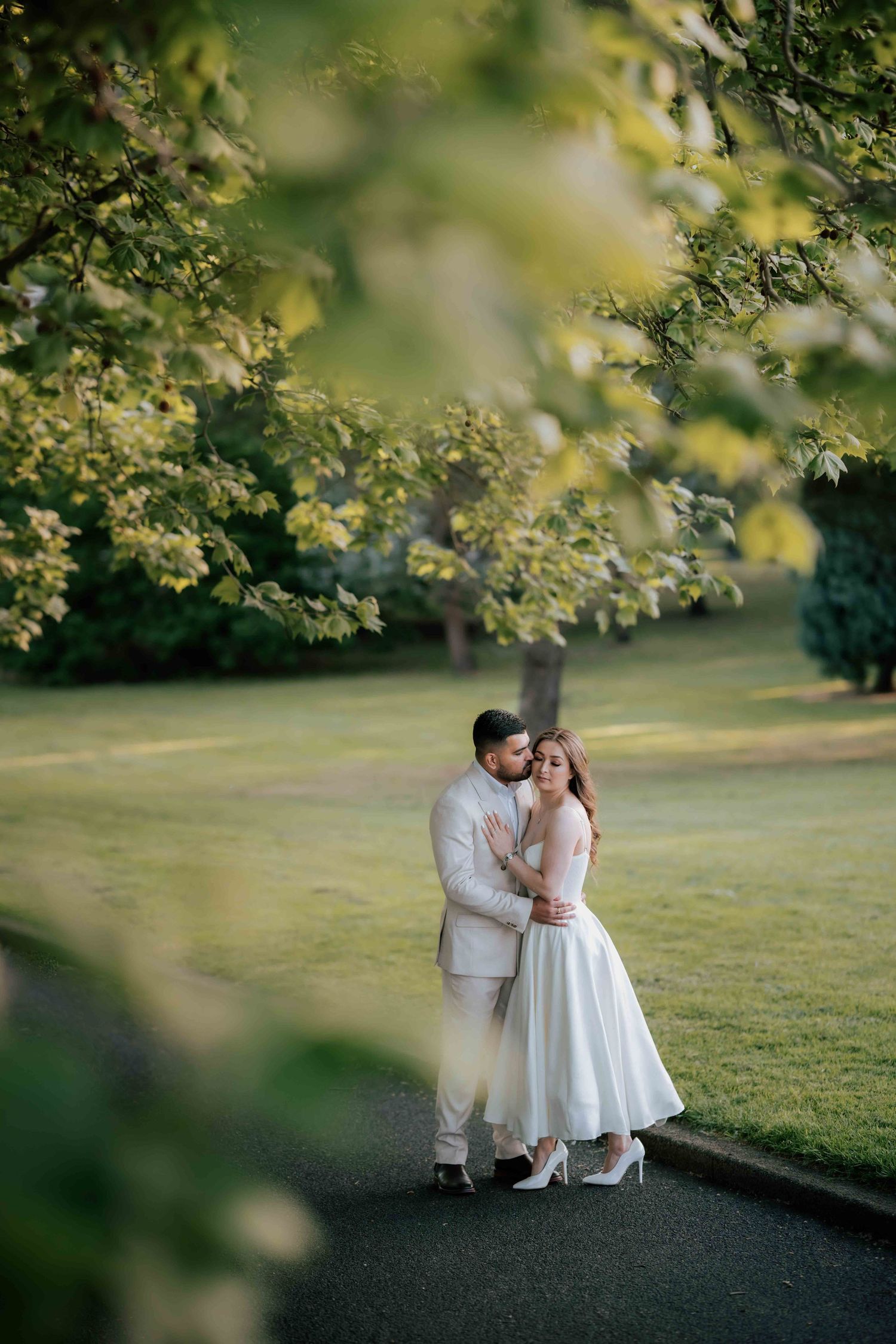 Groom in ivory suit kissing bride on cheek in lush green park framed by leafy branches.