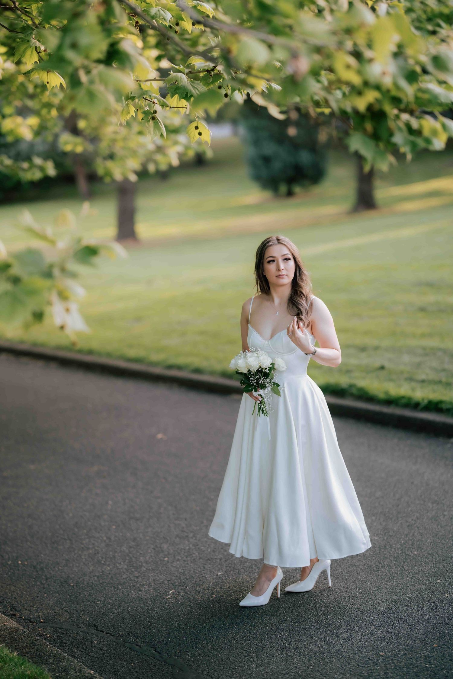 Bride in a white midi dress holding a bouquet while walking along a sunlit park path.