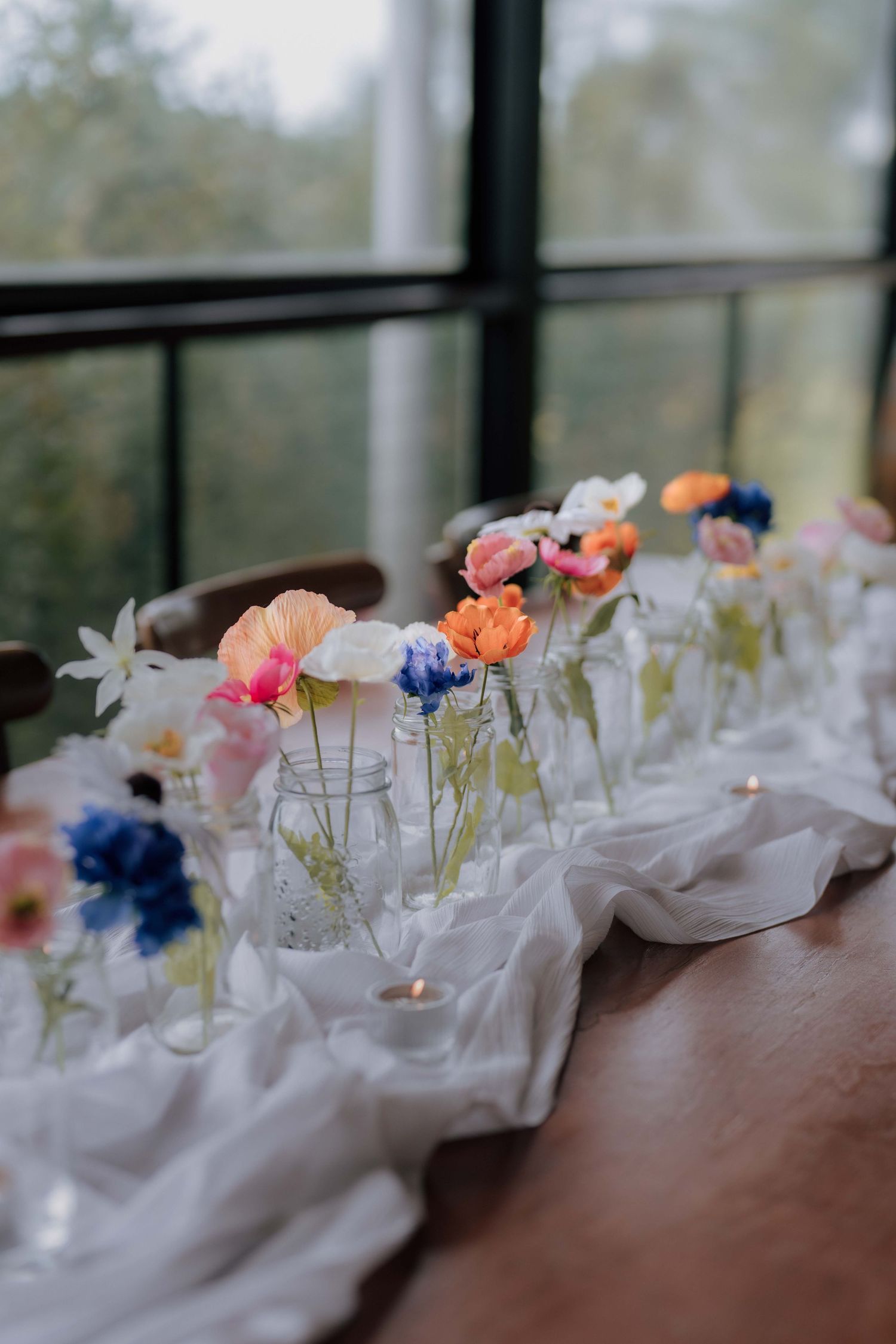 Colorful wildflowers in individual glass bud vases arranged along a white linen table runner with tea lights.