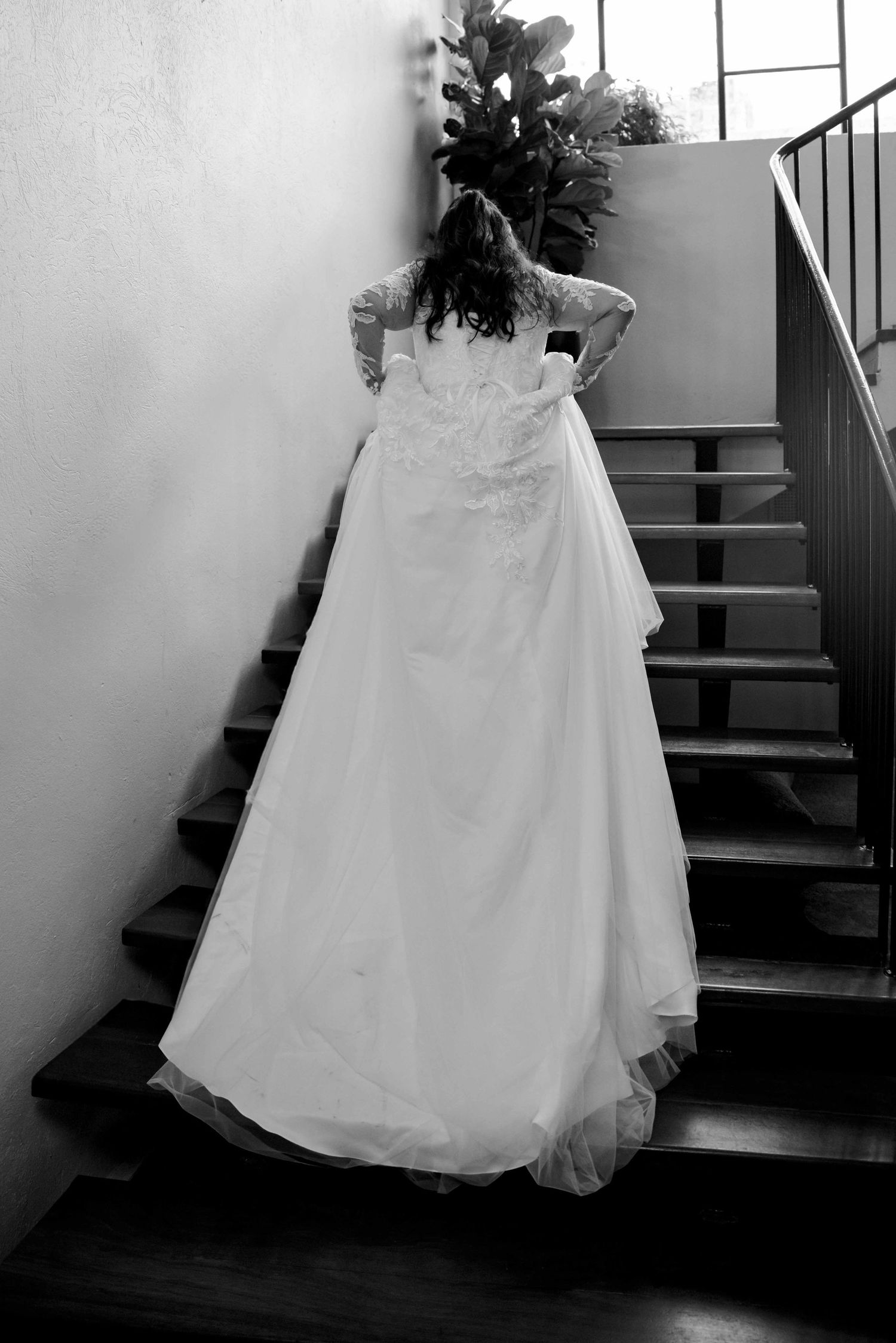 Black and white photo of bride in lace wedding gown standing by staircase with floral bouquet.