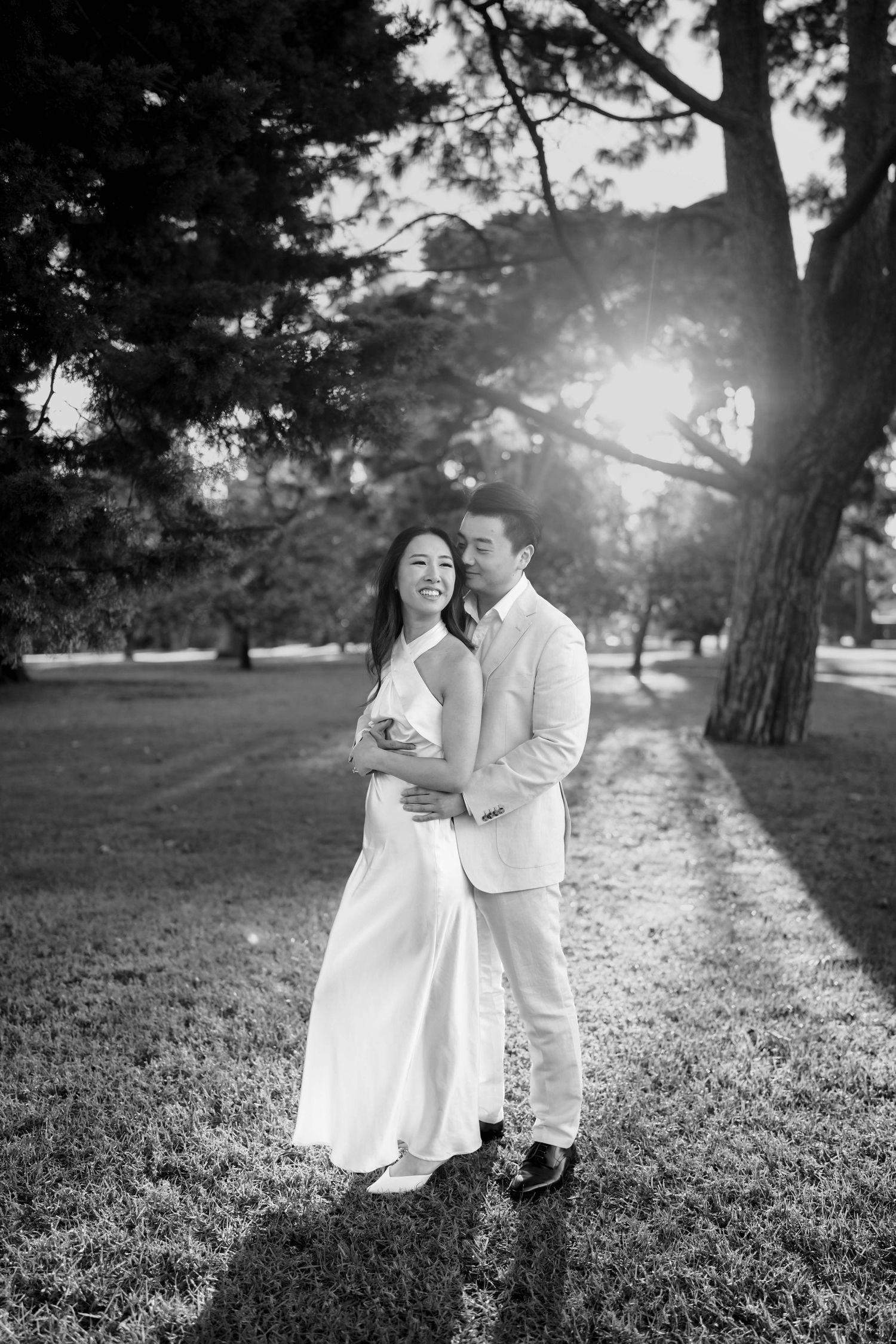 Black and white photo of couple embracing on grass under large trees with sunlight streaming through the branches.