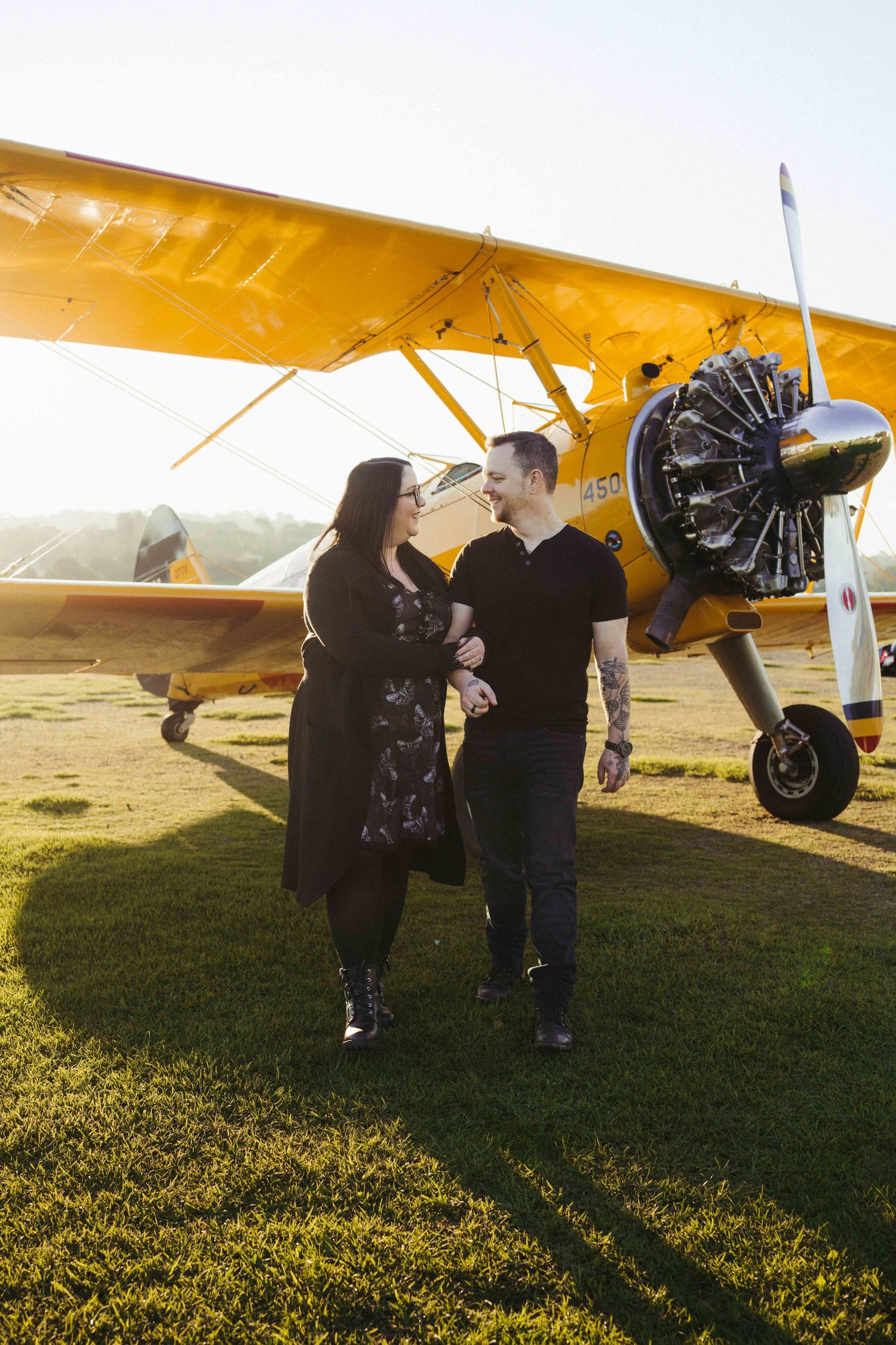 Couple standing together on grass in front of a vintage yellow biplane on a sunny day.