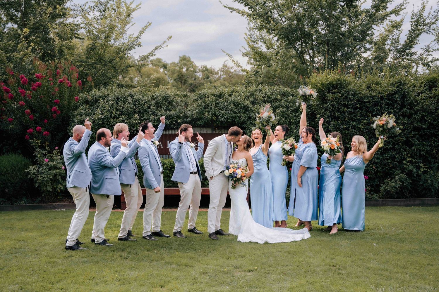 Excited wedding party raises bouquets and cheers around kissing bride and groom outdoors.