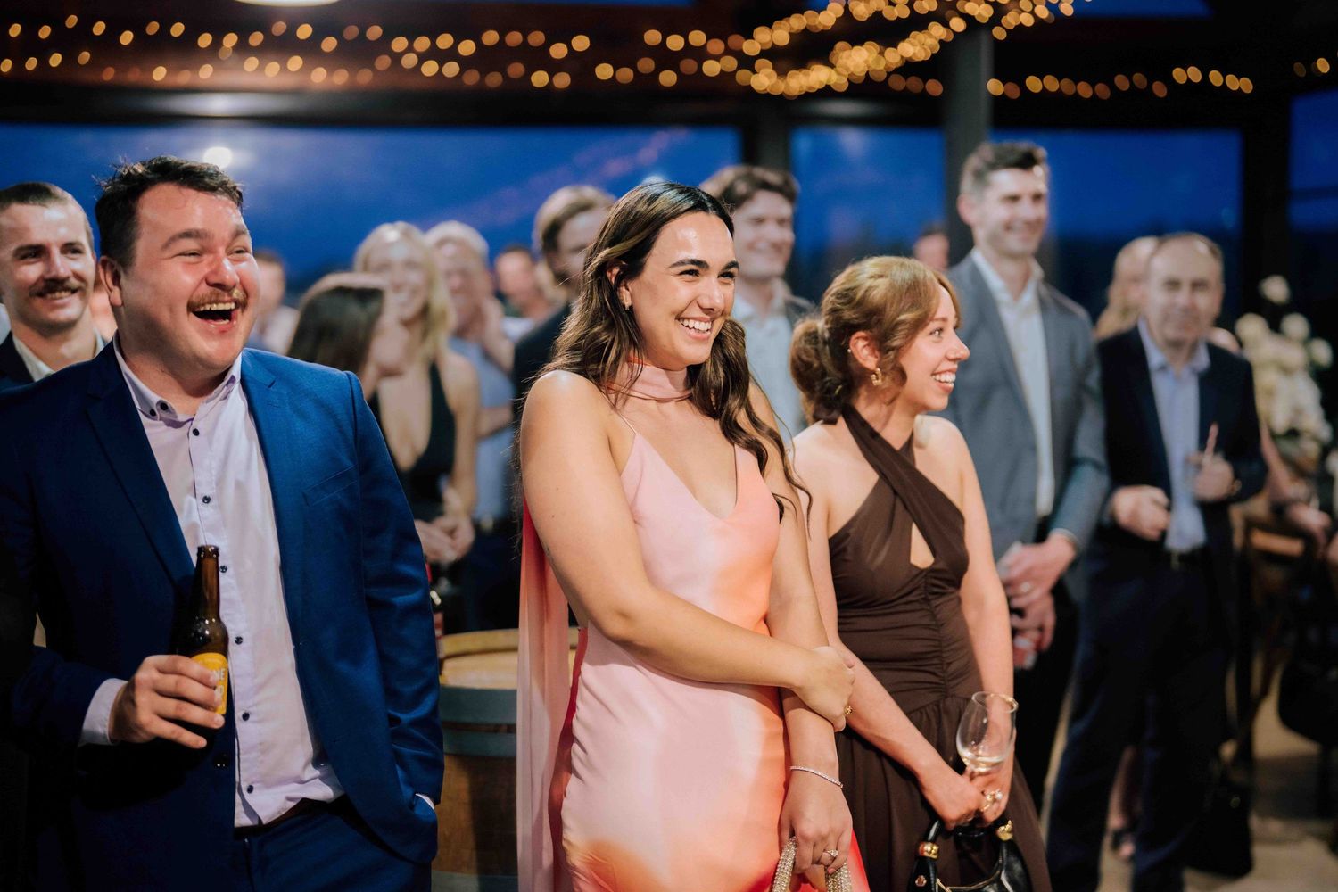 Wedding guests laughing and smiling during a reception celebration with string lights overhead.