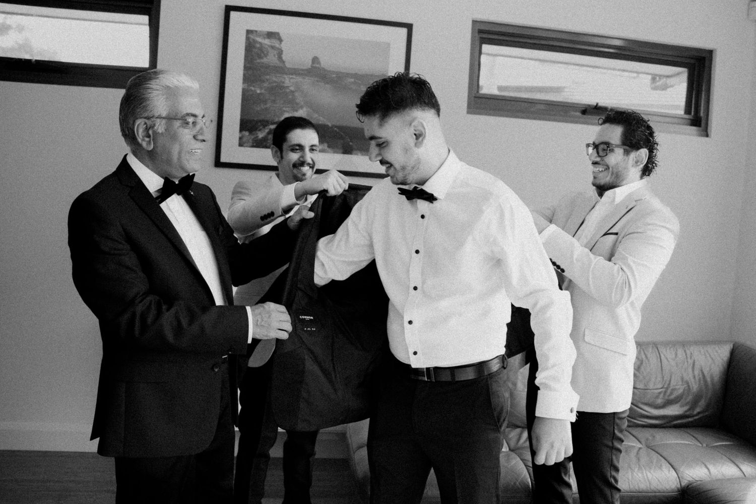 Groom getting bow tie adjusted by father and groomsman before wedding ceremony in black and white photo.