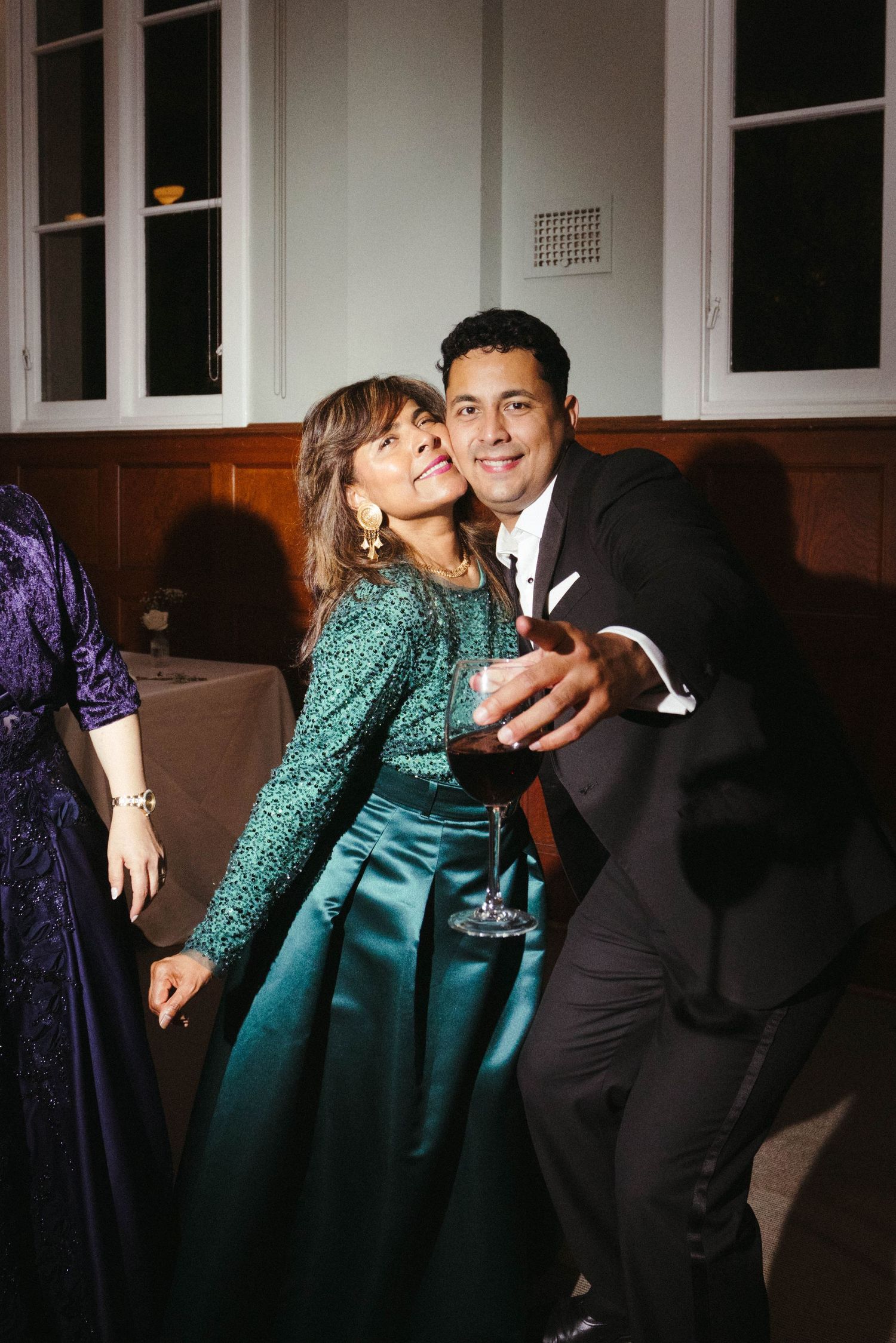 Woman in green sequin gown embracing a man in a black suit holding a wine glass at a formal event.