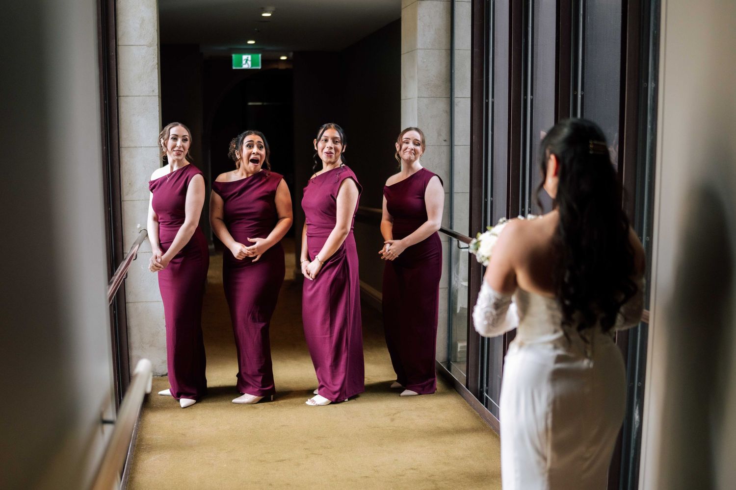 Bride in white gown reveals herself to four bridesmaids in burgundy dresses standing in a hotel corridor.
