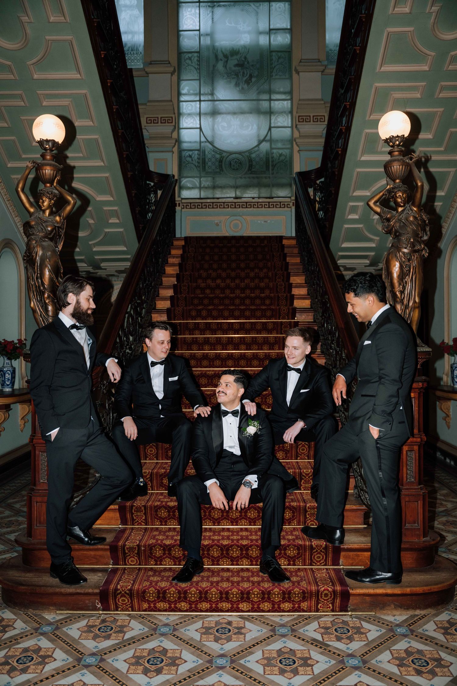Five groomsmen in black tuxedos pose on an ornate staircase with bronze sculptures at an elegant wedding venue.