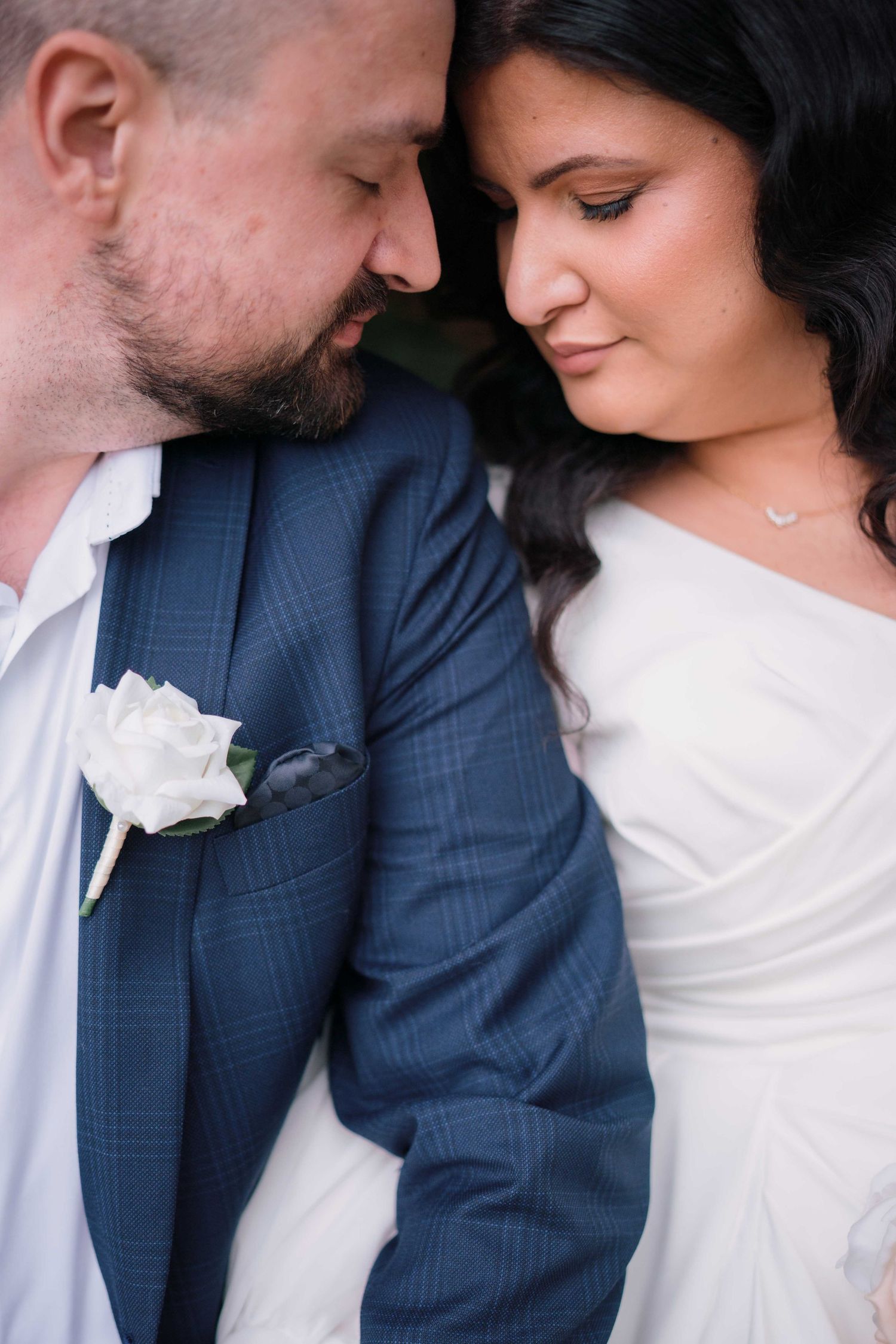 Close-up of bride and groom touching foreheads intimately, groom wearing blue suit with white rose boutonniere.