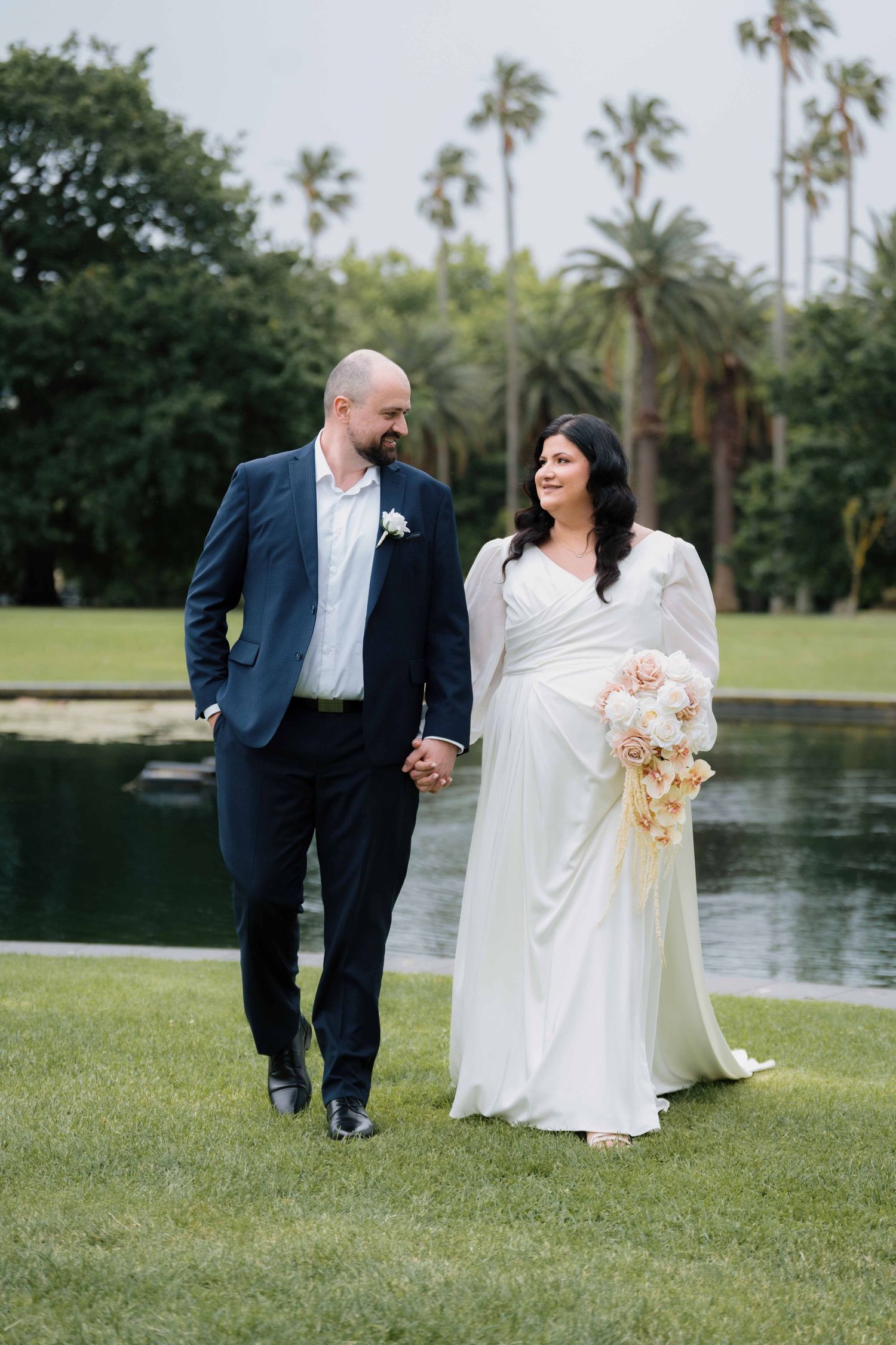 Bride and groom walk hand in hand beside a serene pond surrounded by tall palm trees.
