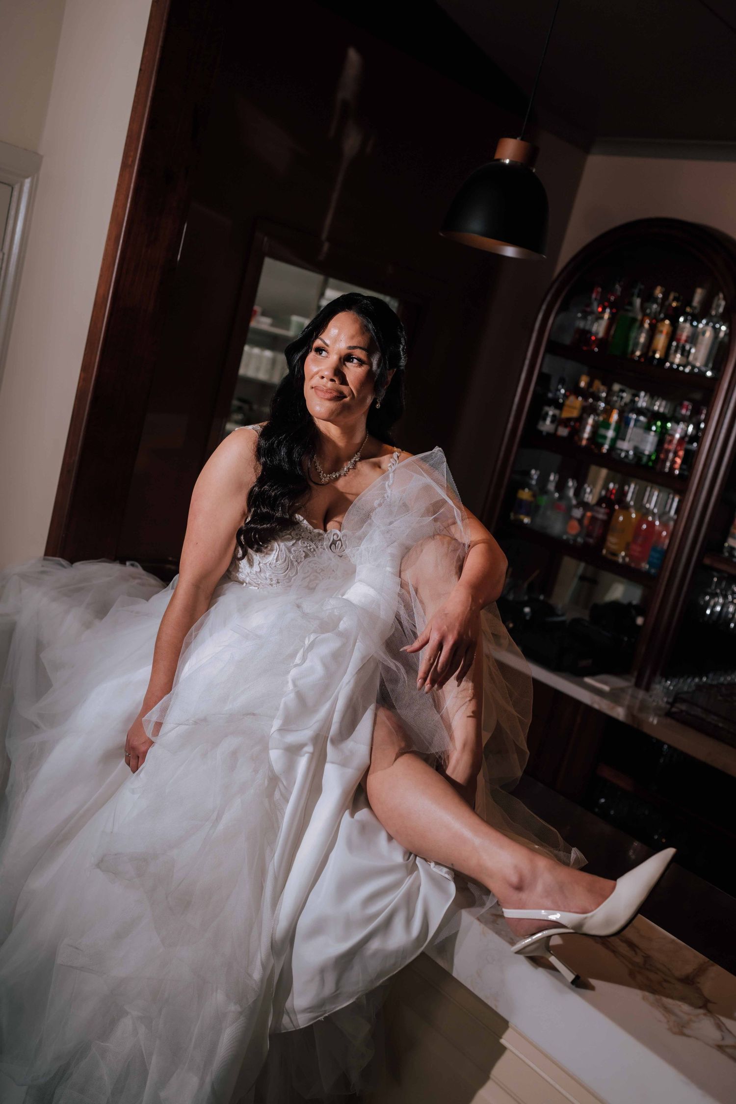 A bride in a white wedding gown shows her heels while seated at a bar in a dimly lit venue.