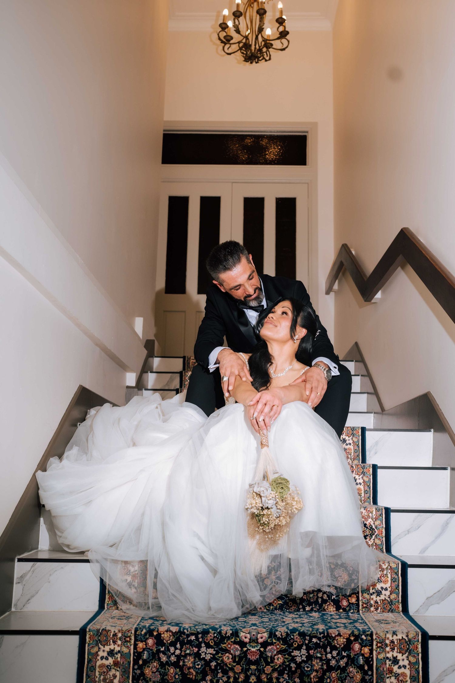 Bride and groom sitting together on marble staircase, sharing a romantic moment with bouquet.