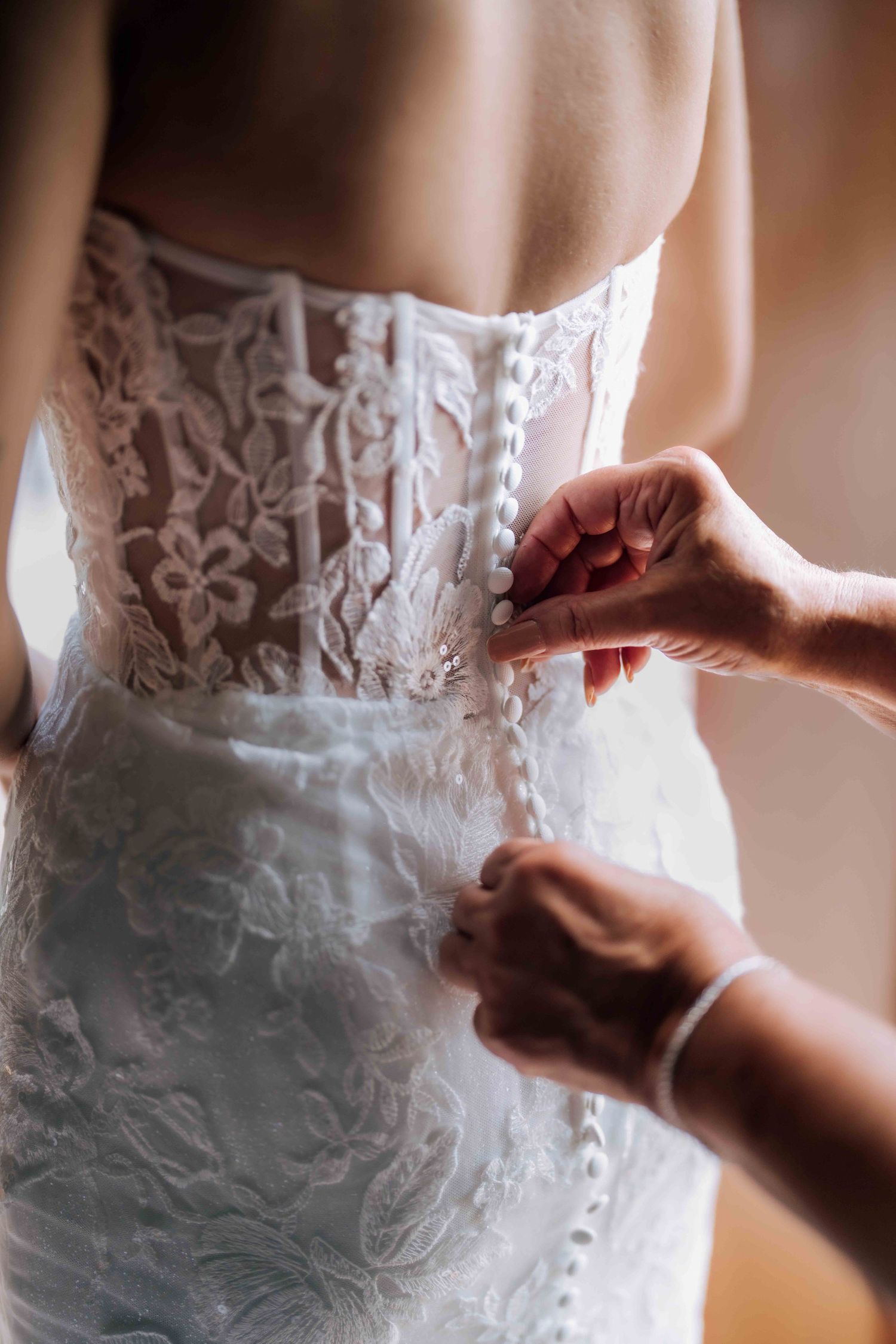 Close-up of hands buttoning the back of a white lace wedding dress with pearl buttons.