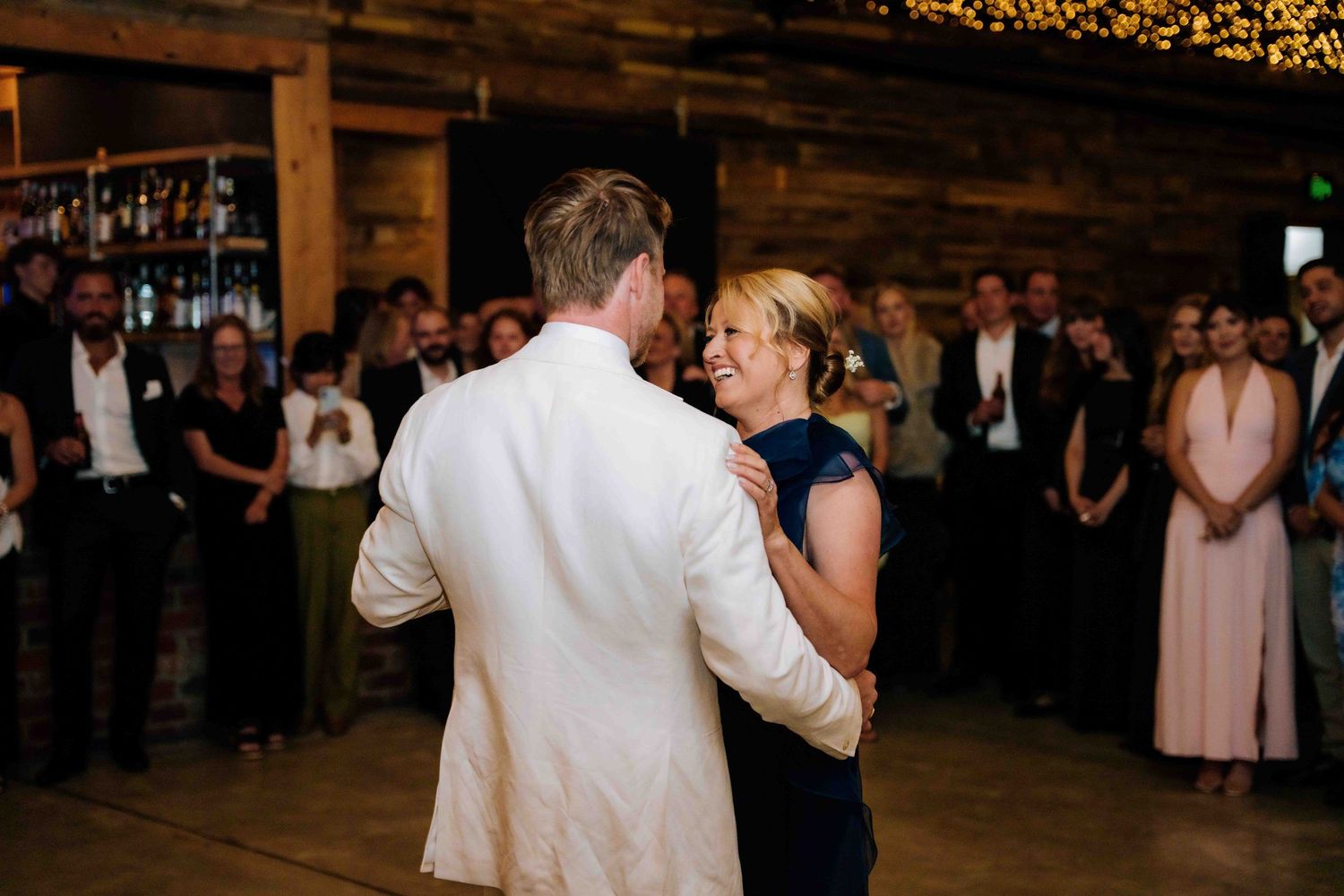 Groom in white suit shares a dance with a woman in navy dress at rustic barn wedding reception.