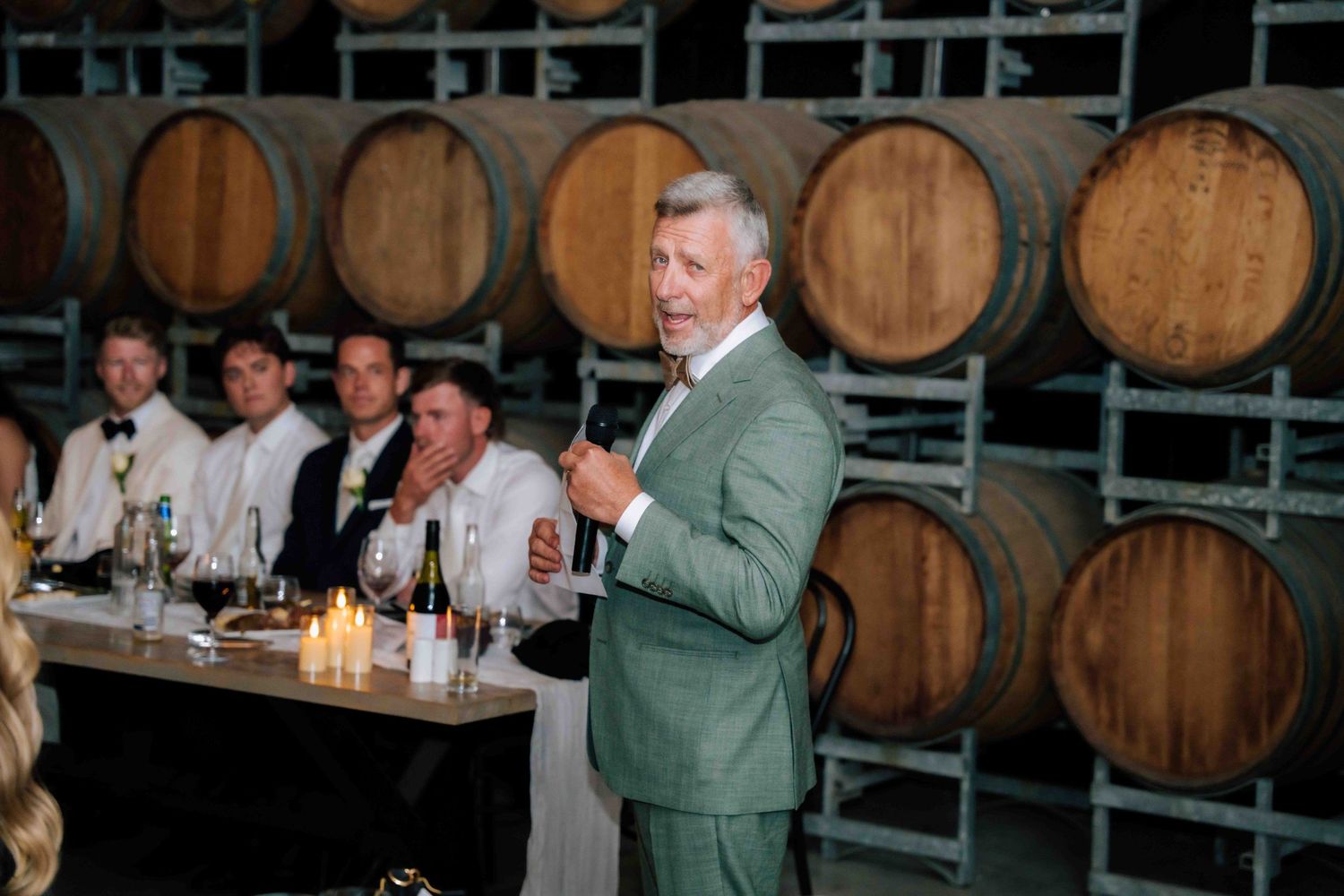 Man in green suit gives speech beside wine barrels at wedding reception.