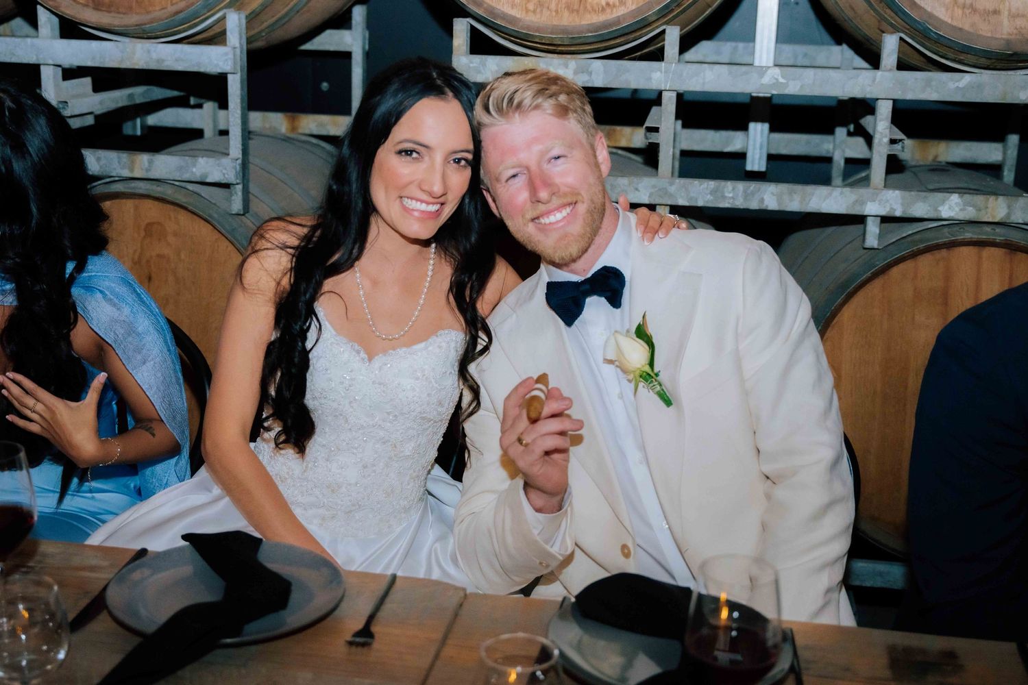 Bride and groom smile together at wedding reception table with wine barrels in background.