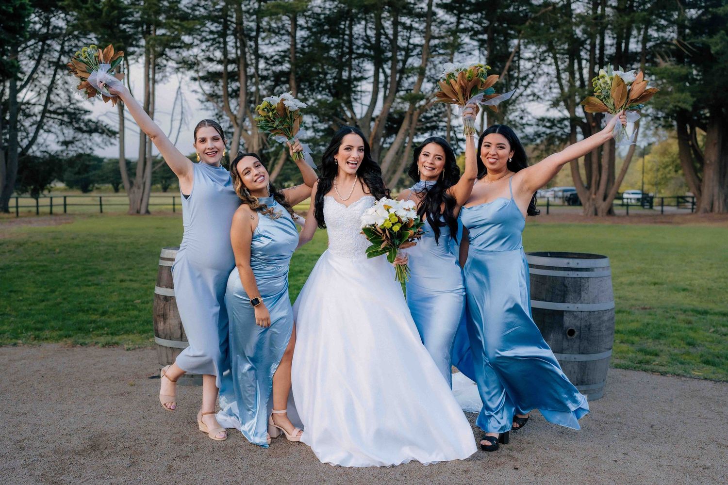 Bride in white gown celebrates with four bridesmaids in blue satin dresses, raising bouquets outdoors near barrels.