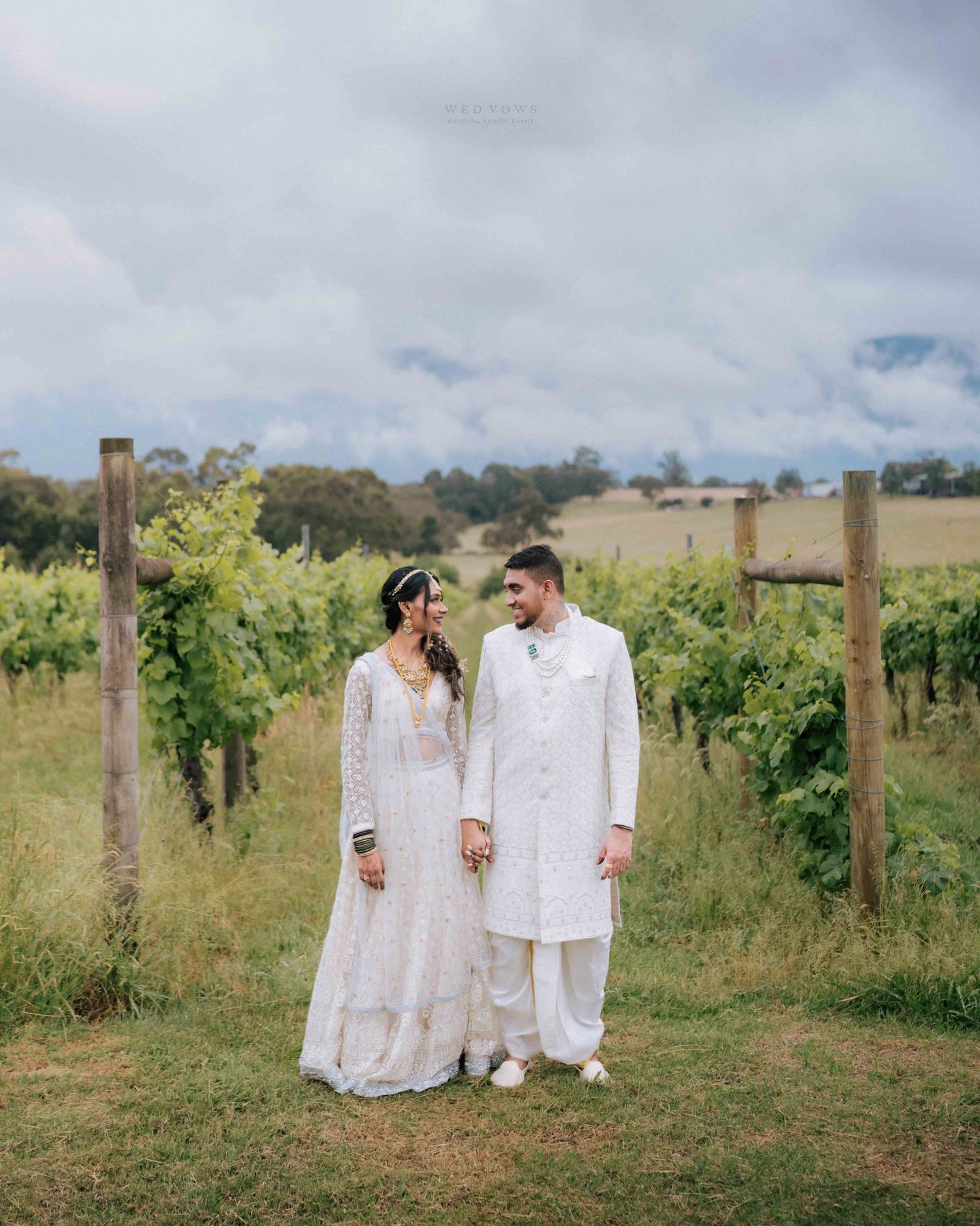 Indian couple in white traditional wedding attire share a loving gaze in a vineyard setting.