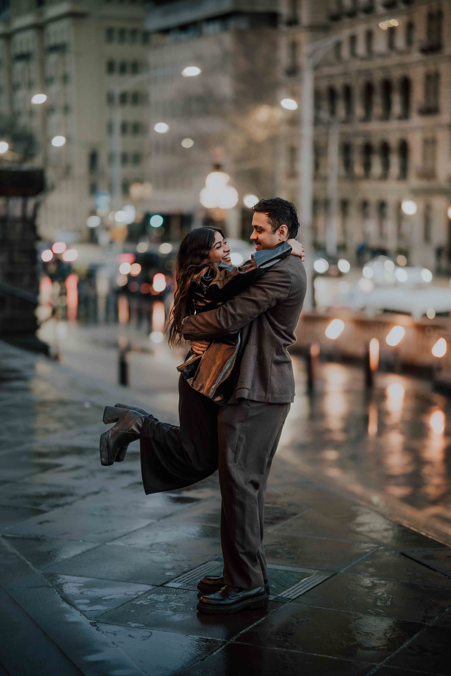 Man lifting woman in a joyful embrace on a rainy city street at night with bokeh lights.