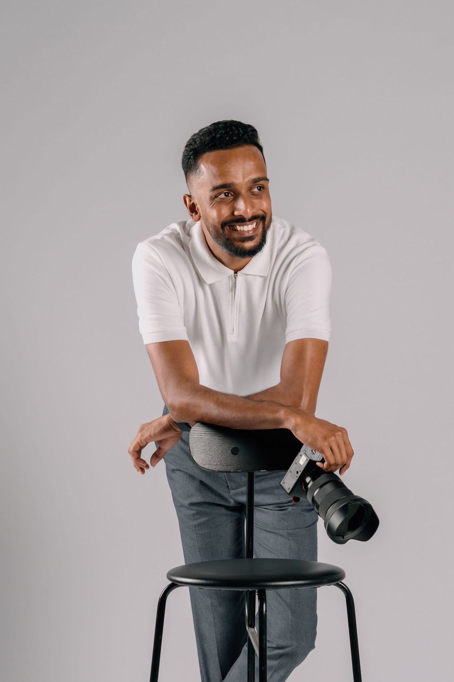 Smiling male photographer leaning on black stool holding DSLR camera in white polo shirt.