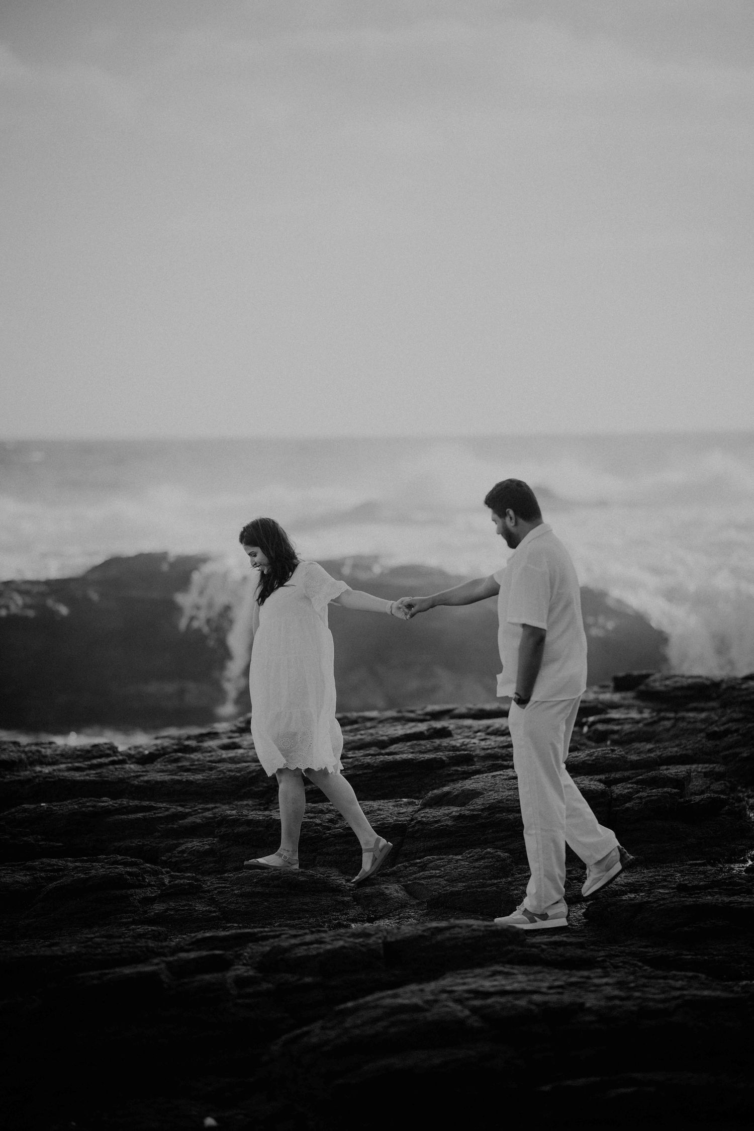 Black and white photo of couple holding hands while walking on rocky coastal terrain.