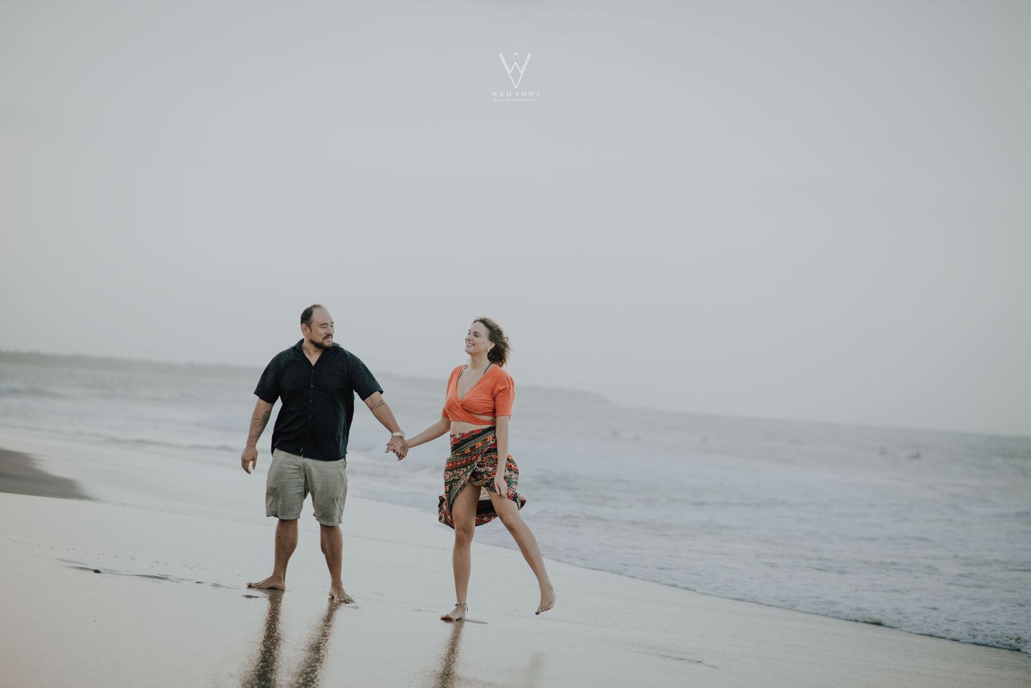 Couple holding hands and walking along a sandy beach with ocean waves behind them.