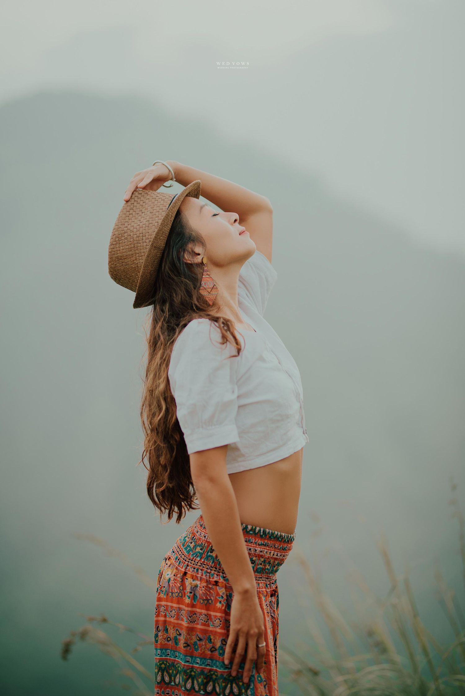 Woman in boho outfit holding straw hat while posing against misty mountain backdrop.