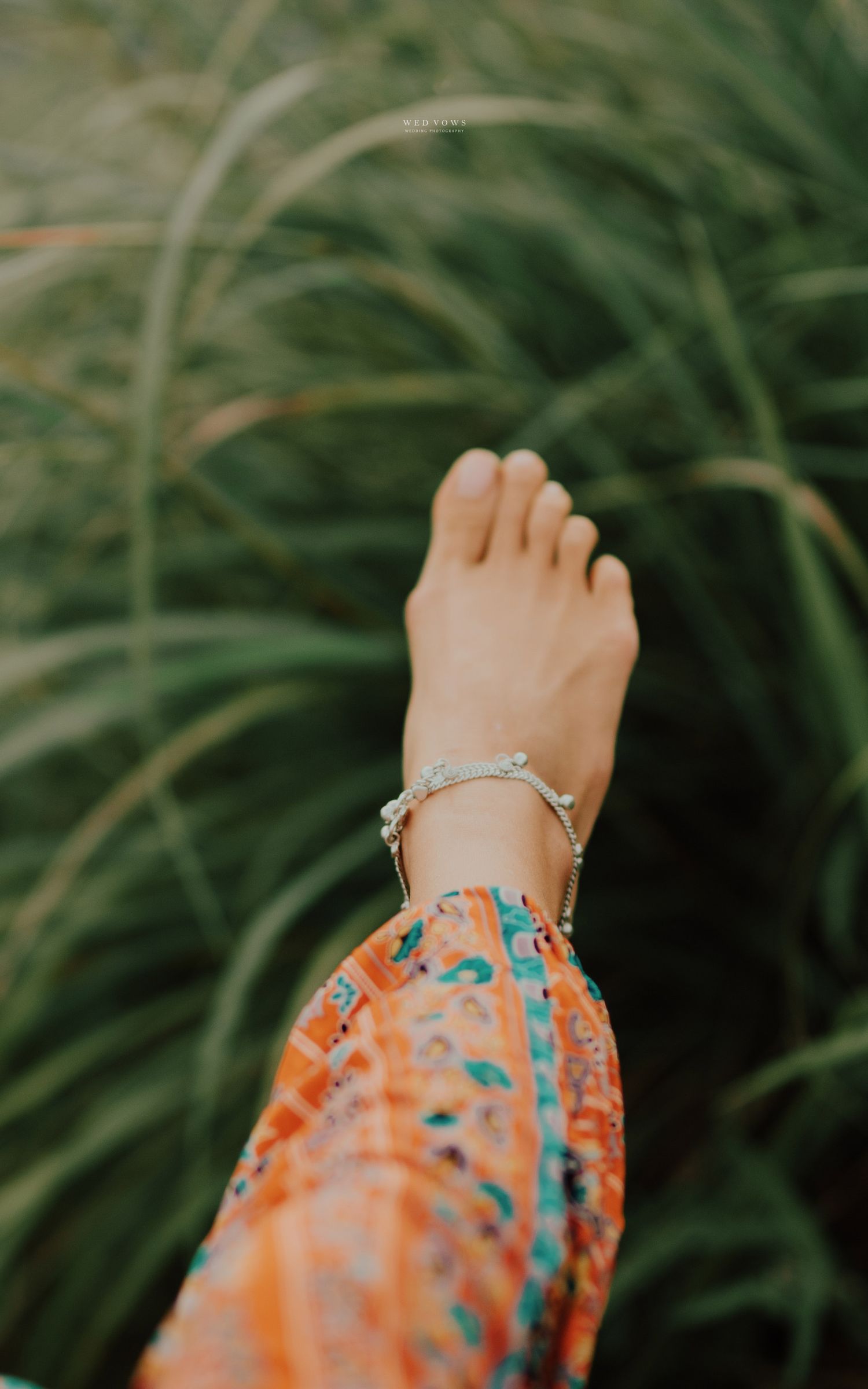 A bare foot with a delicate silver anklet raised against lush green grass, wearing a colorful boho orange floral skirt.