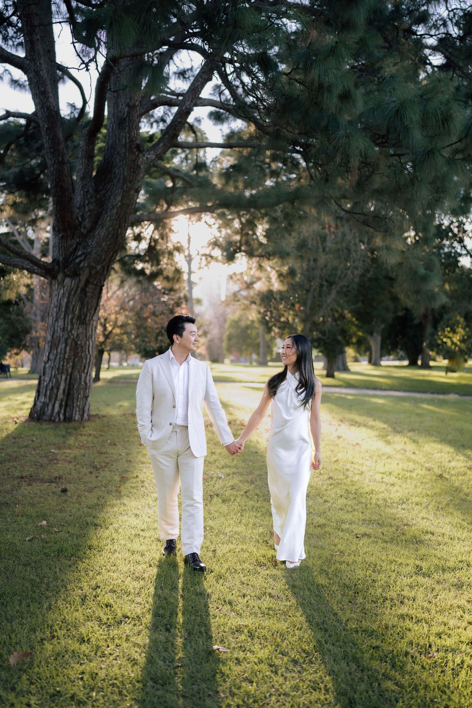 Couple in white wedding attire holding hands and walking through a sunlit park with tall green trees.